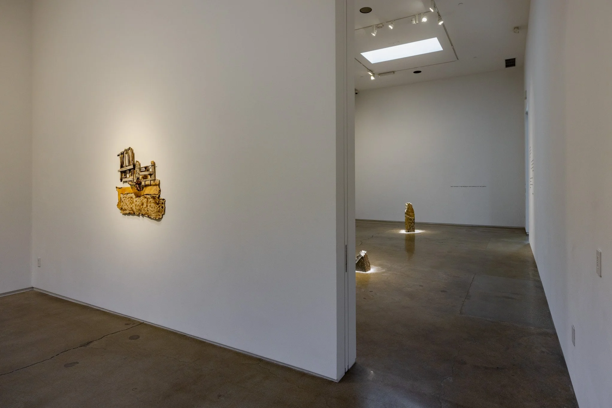 Contemporary art gallery with minimalist white walls and sculptures, including a small mixed-media piece on the left wall and two illuminated stone sculptures on the floor in the background, with skylights overhead.