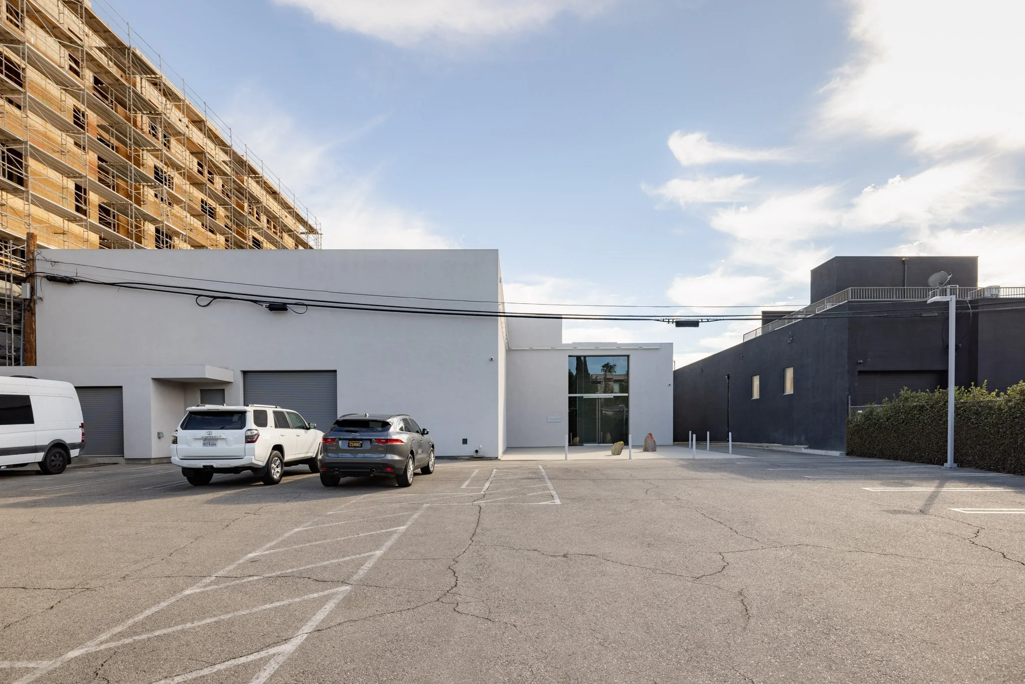Empty parking lot with three parked cars in front of a modern white and black building, under a partly cloudy sky.