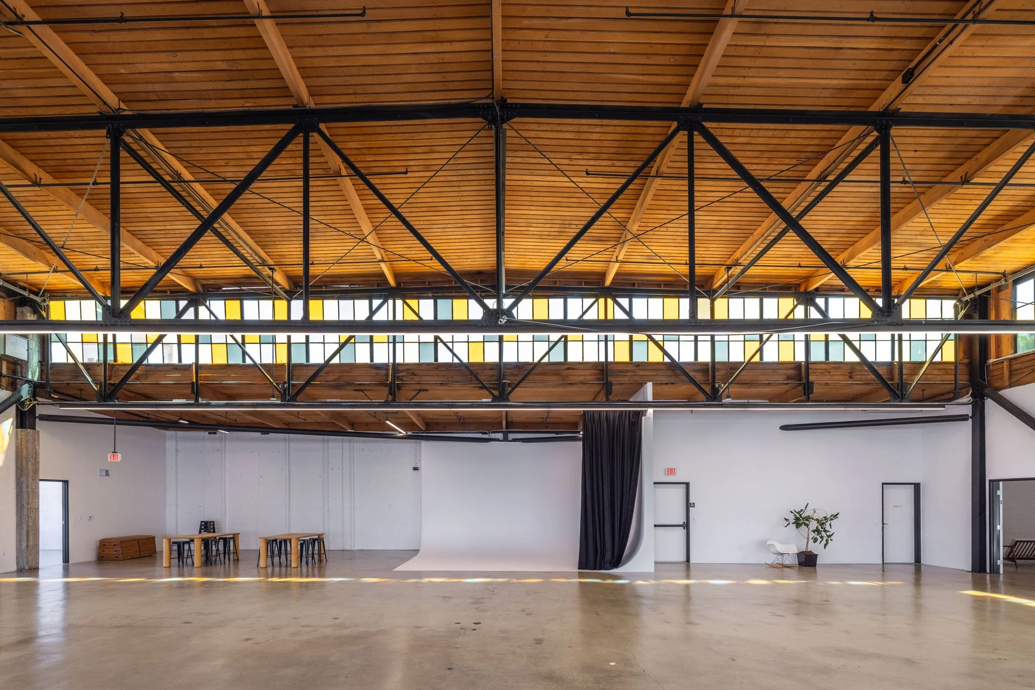 Spacious, industrial-style multipurpose event space with wooden ceiling, black metal beams, white walls, a small table and chairs, a potted plant, a black curtain, and large windows near the ceiling.