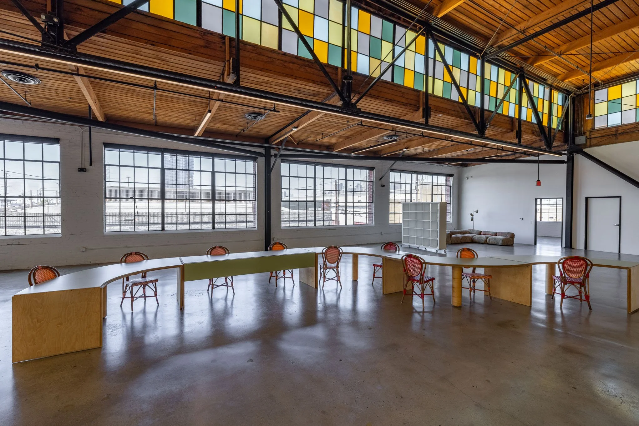 Spacious industrial-style room with large windows, wooden ceiling, and colorful stained glass. Contains a curved table surrounded by red chairs, with a white shelving unit and a lounge area with cushions in the background.