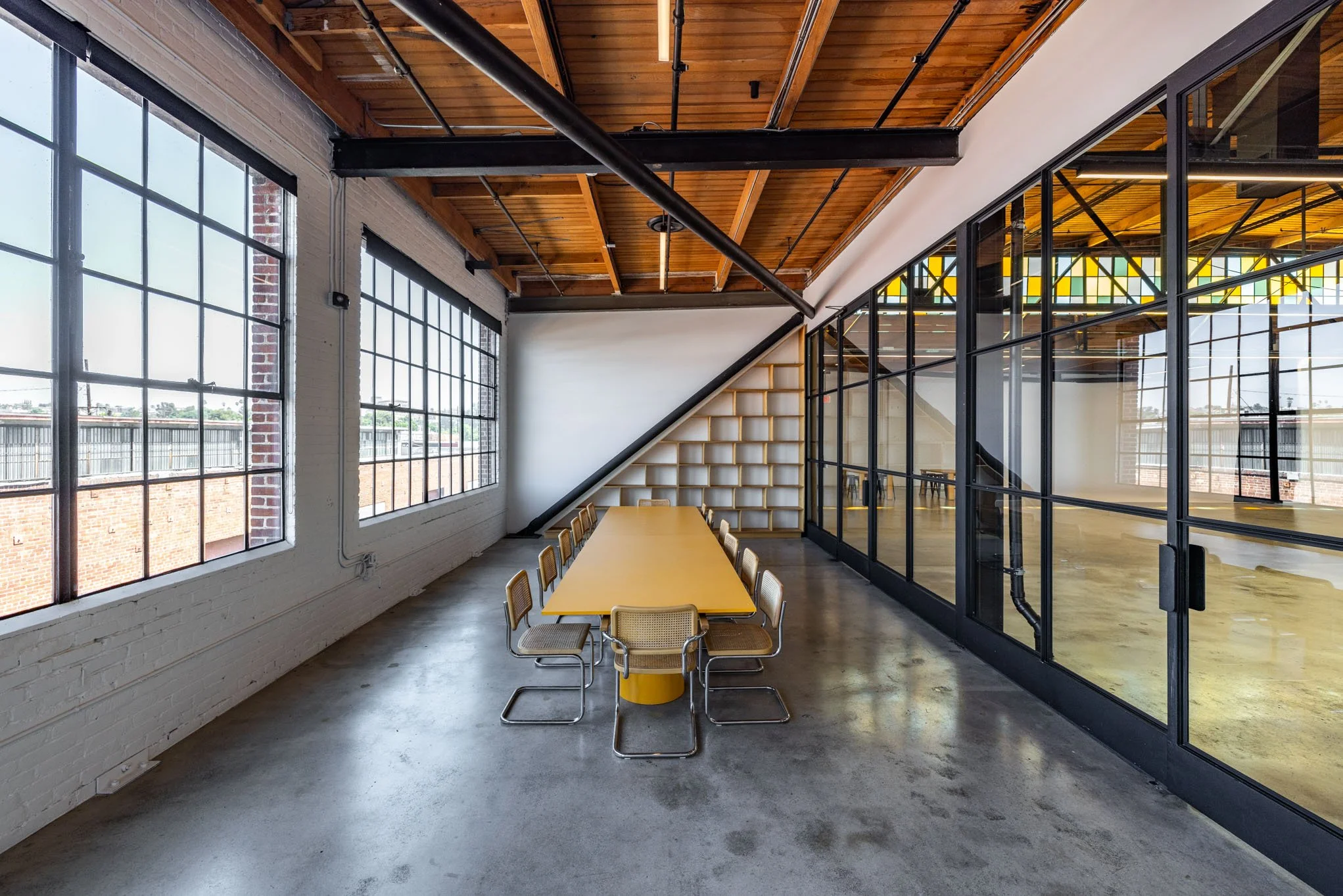 A modern conference room with large industrial-style windows, a yellow table surrounded by chairs, and a glass wall with black framing separating it from an open space.