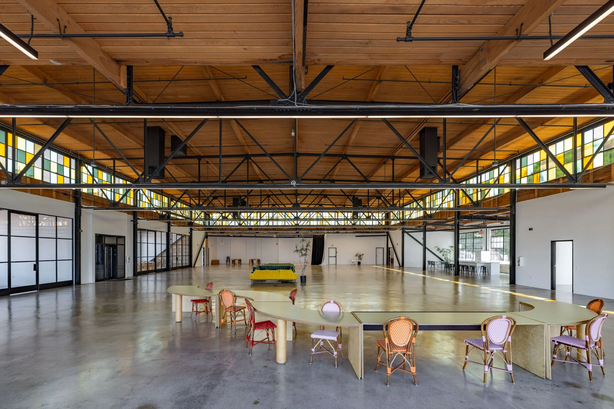 Interior view of a spacious, modern event hall with a high wooden ceiling, stained glass windows, a long curved table with colorful chairs, a yellow and green sofa, and potted plants.