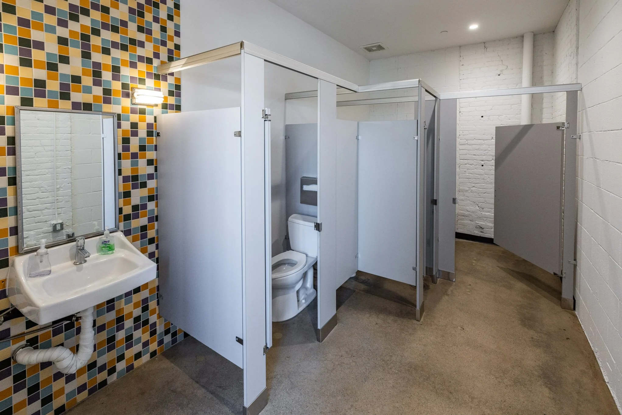 Public restroom with multiple stalls, a small sink with soap and hand sanitizer, and colorful tile wall.