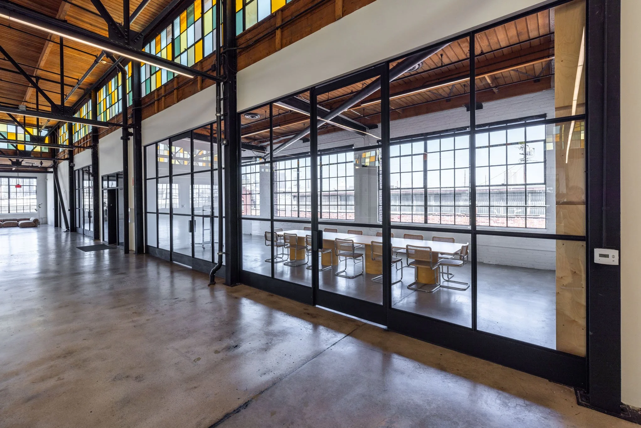 Interior of a modern industrial-style office space with large windows, exposed wooden ceiling, and glass partitioned meeting room with tables and chairs.