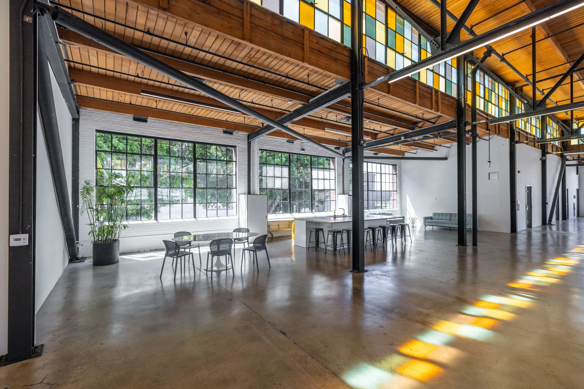 Open industrial-style loft with large windows, wooden and metal ceiling, and scattered seating including a potted plant, tables, chairs, and a gray sofa.