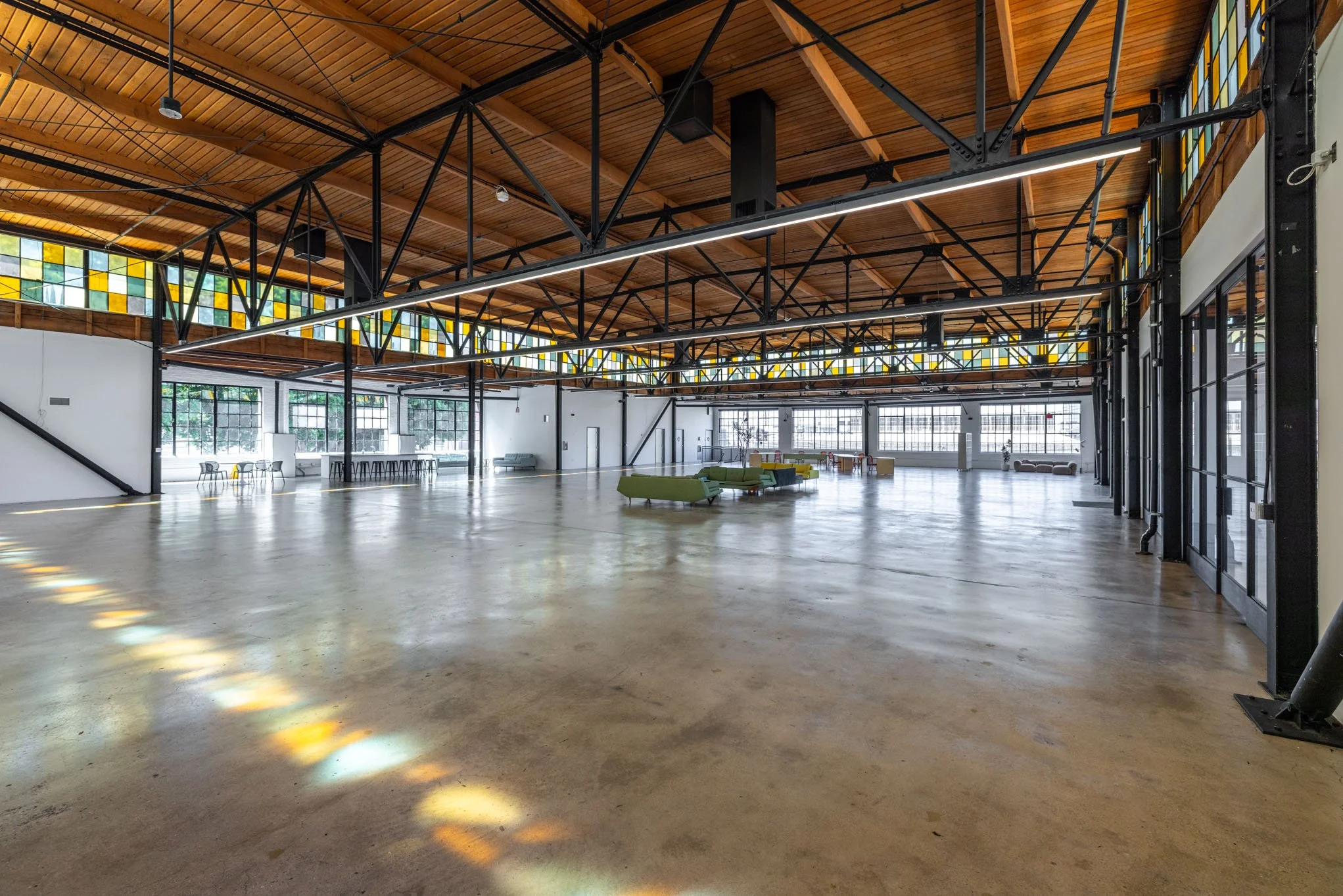 Large, empty industrial-style interior space with wooden ceiling, black metal beams, concrete floor, and large windows with stained glass accents.