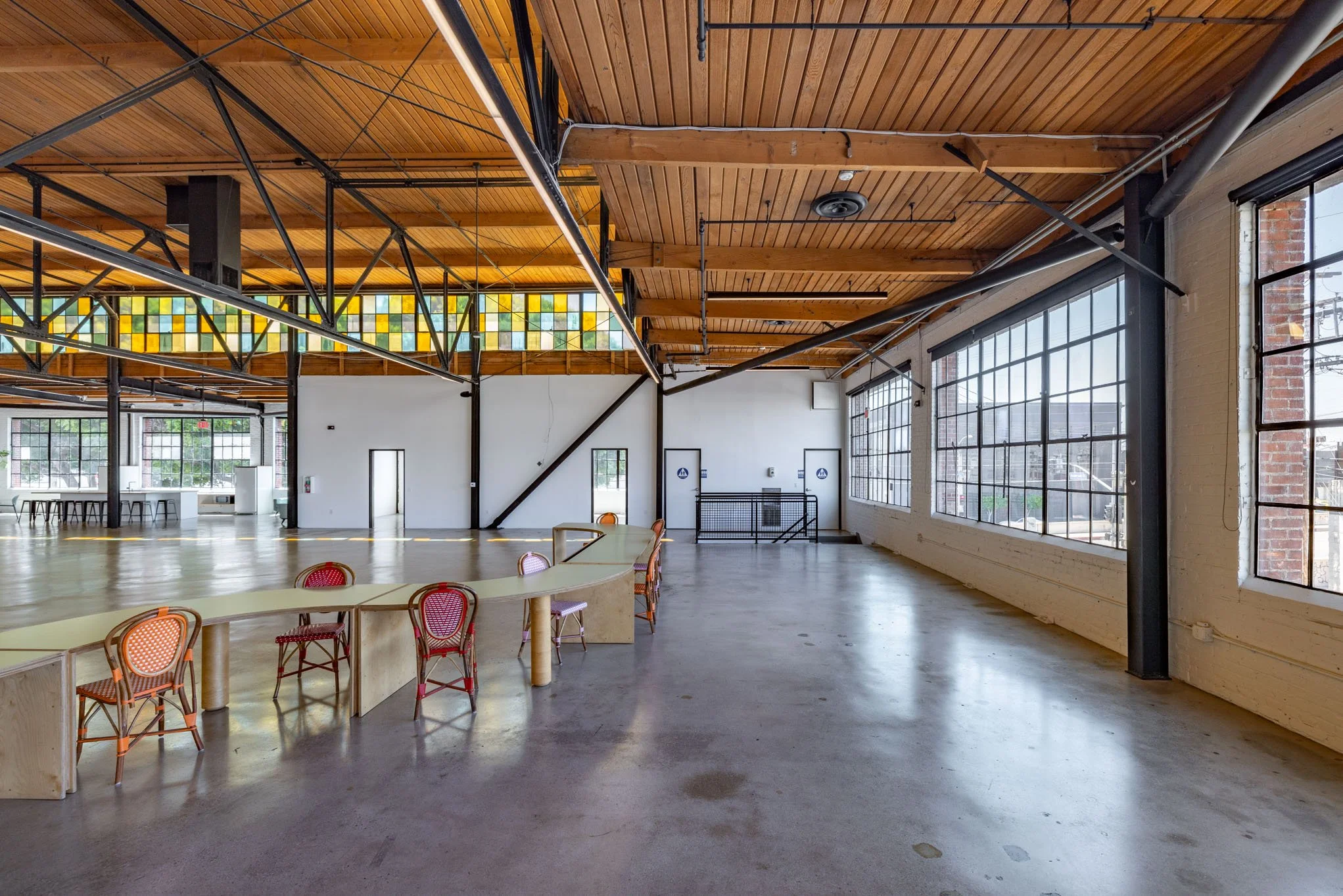 Large open indoor space with high wooden ceiling, metal beams, large windows, and minimal furniture, including a curved table with chairs. Bright natural light fills the room.