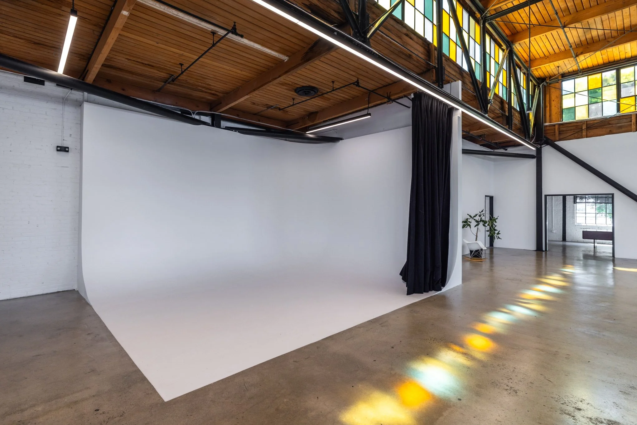 Photography studio with a white seamless backdrop, black curtains, and industrial-style ceiling with wooden beams and stained glass windows.