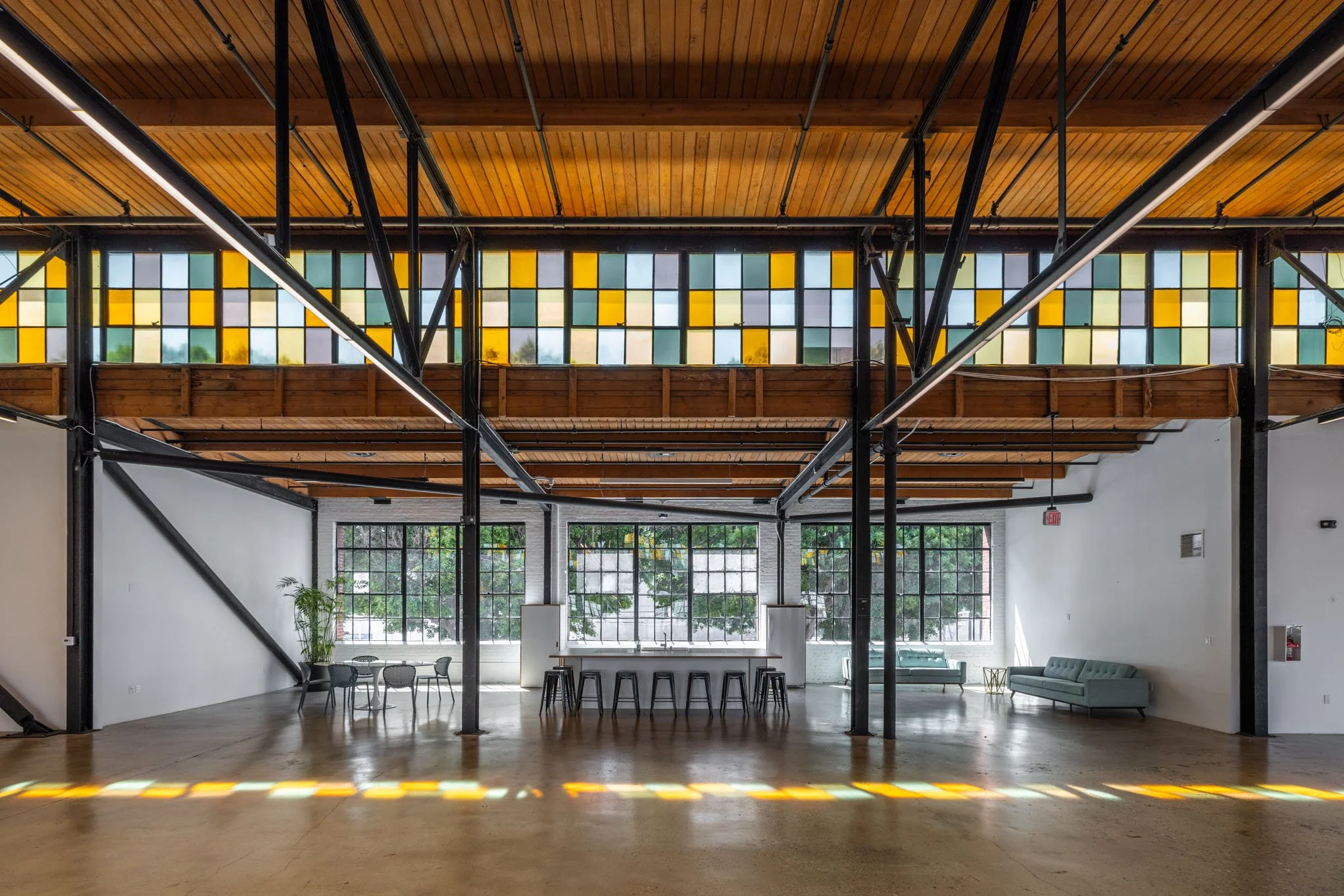 Interior of a modern industrial-style building with large windows, colorful stained glass ceiling, wooden ceiling panels, black metal beams, and minimalist furniture.
