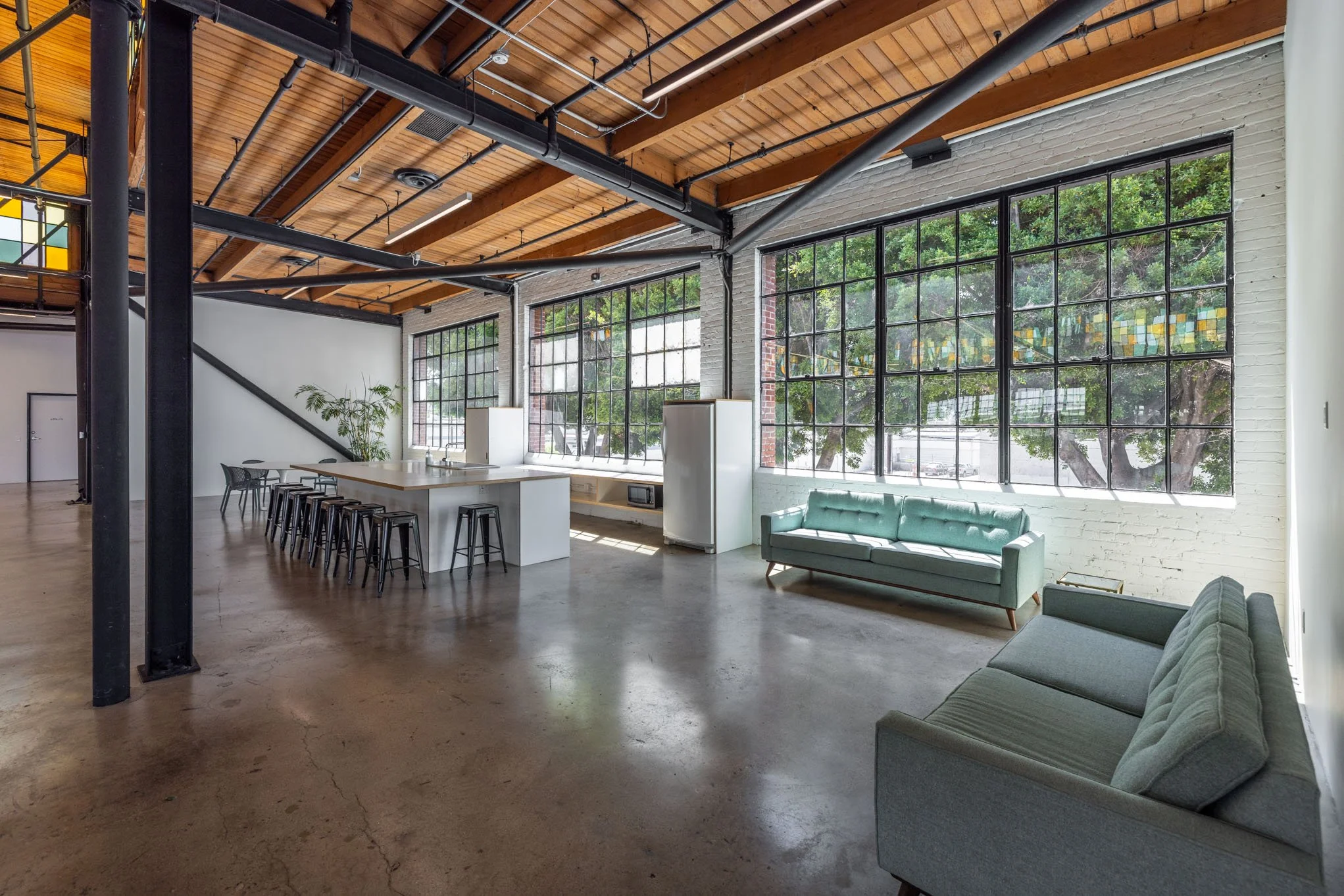 Spacious modern loft with large grid windows, wooden ceiling, polished concrete floors, and minimalist furniture including a gray sofa, teal loveseat, and a white kitchen island with black stools.