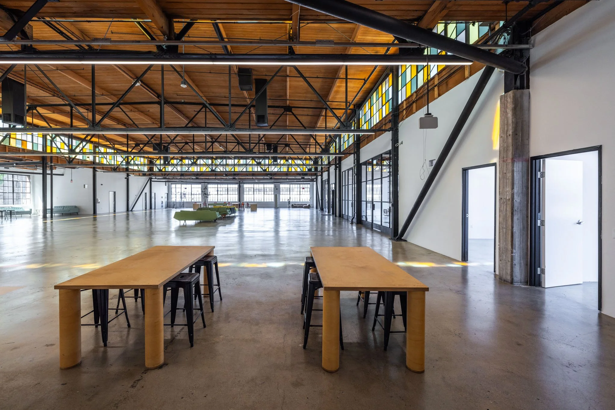 Interior of a spacious, industrial-style event hall with high wooden ceilings, black metal beams, large windows with stained glass accents, and a polished concrete floor. There are two wooden tables with black stools in the foreground and some seatin