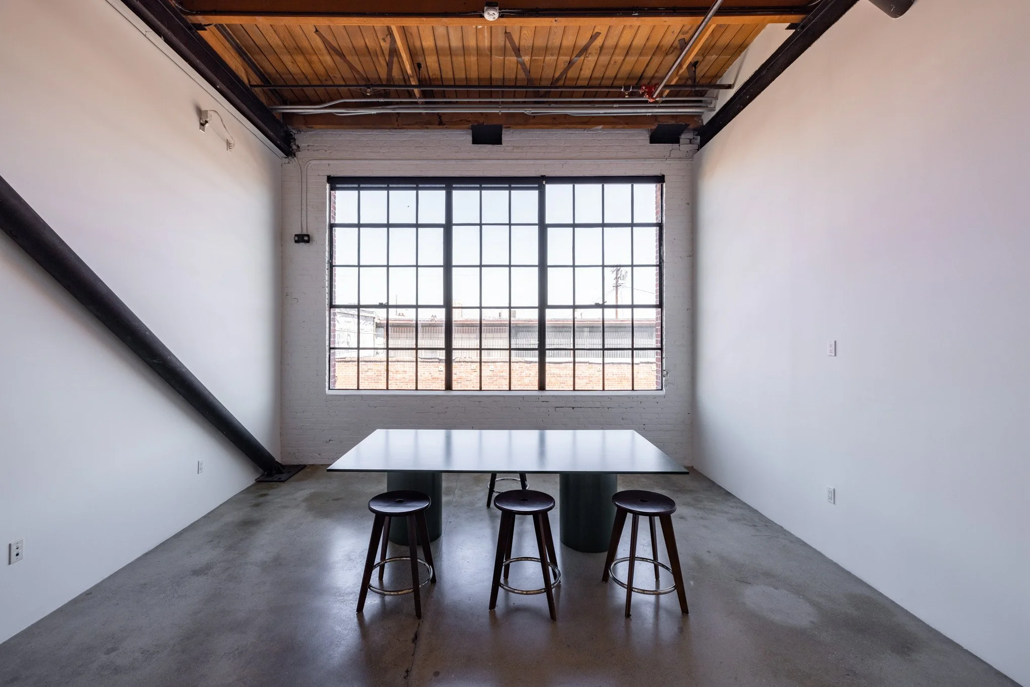 Empty room with white walls, a large window with multiple panes, a black pipe on the left wall, wooden ceiling, concrete floor, and a rectangular table with three stools underneath.