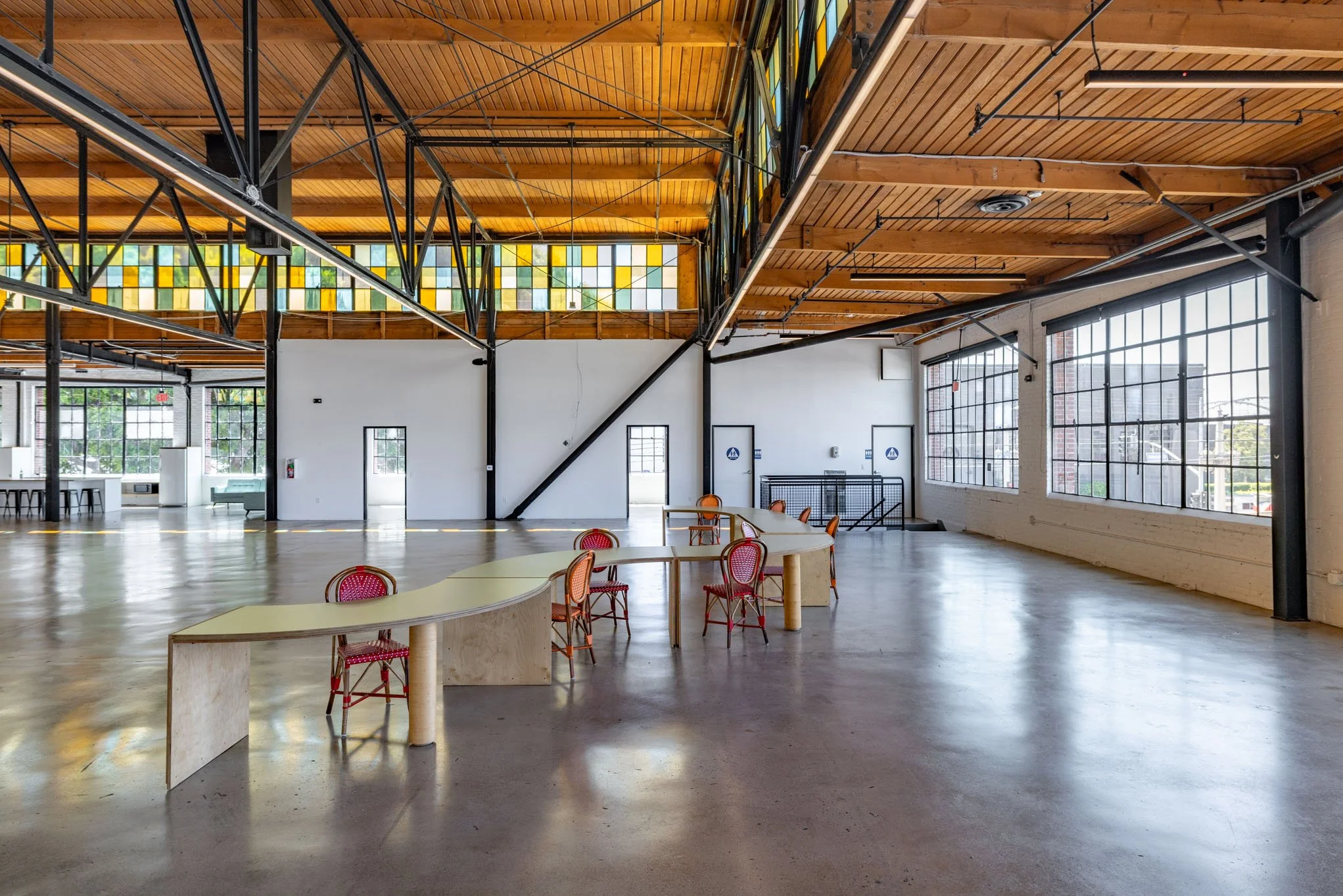 Empty industrial-style event space with large windows, wooden ceiling, and a few tables and chairs.