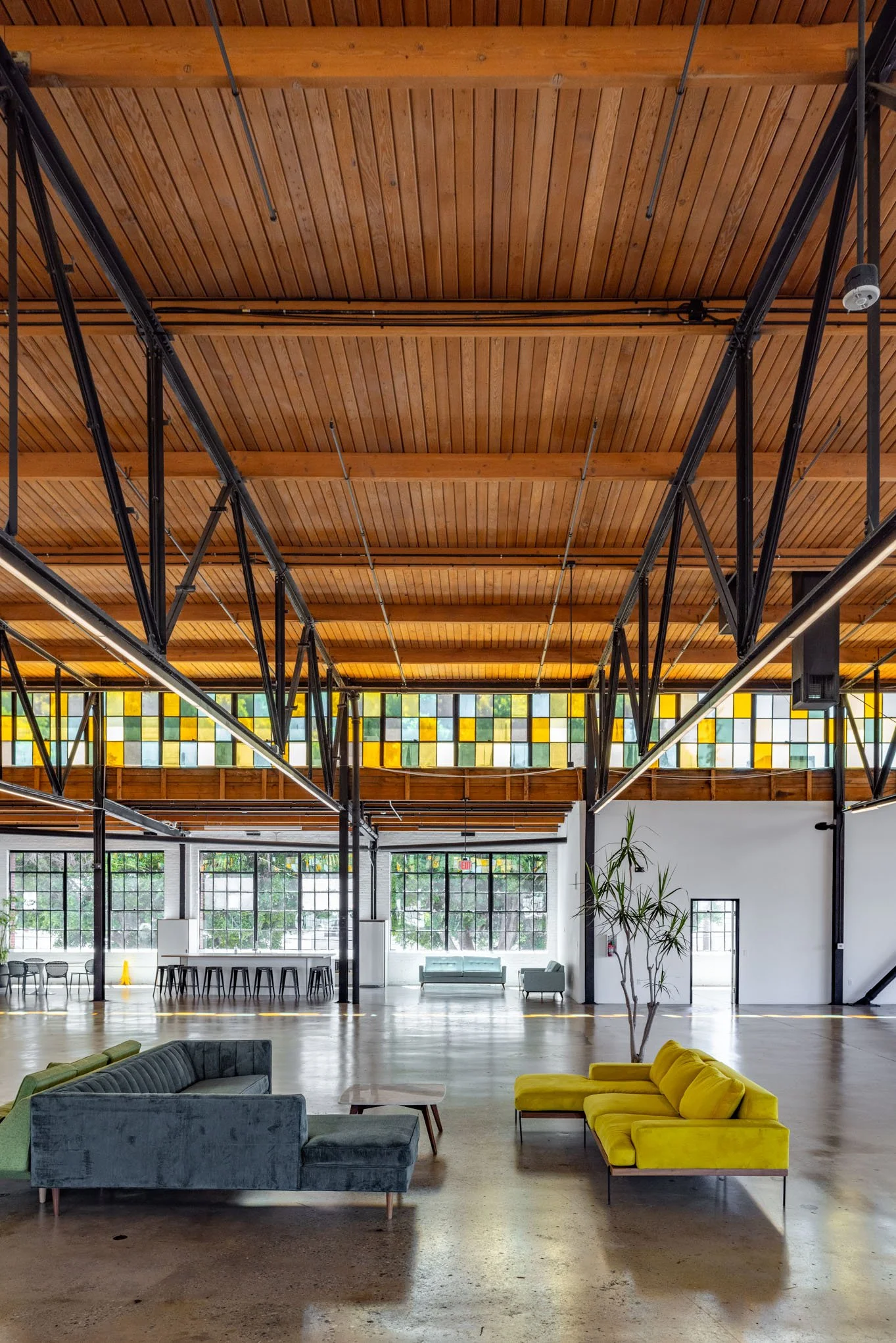Interior of a spacious, industrial-style lounge with high wooden ceilings, large windows, and colorful stained glass accents. There are modern sofas in green and yellow, a few armchairs, small tables, and some plants.