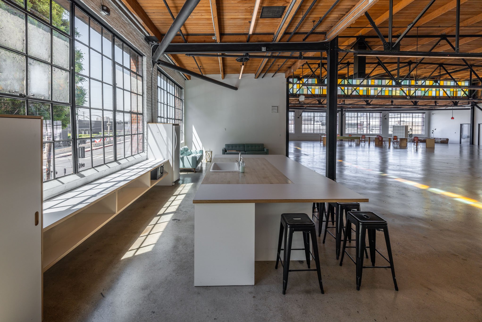 Interior of a spacious industrial style open-concept room with large windows, a kitchen island with stools, seating area, and views of an empty floor.