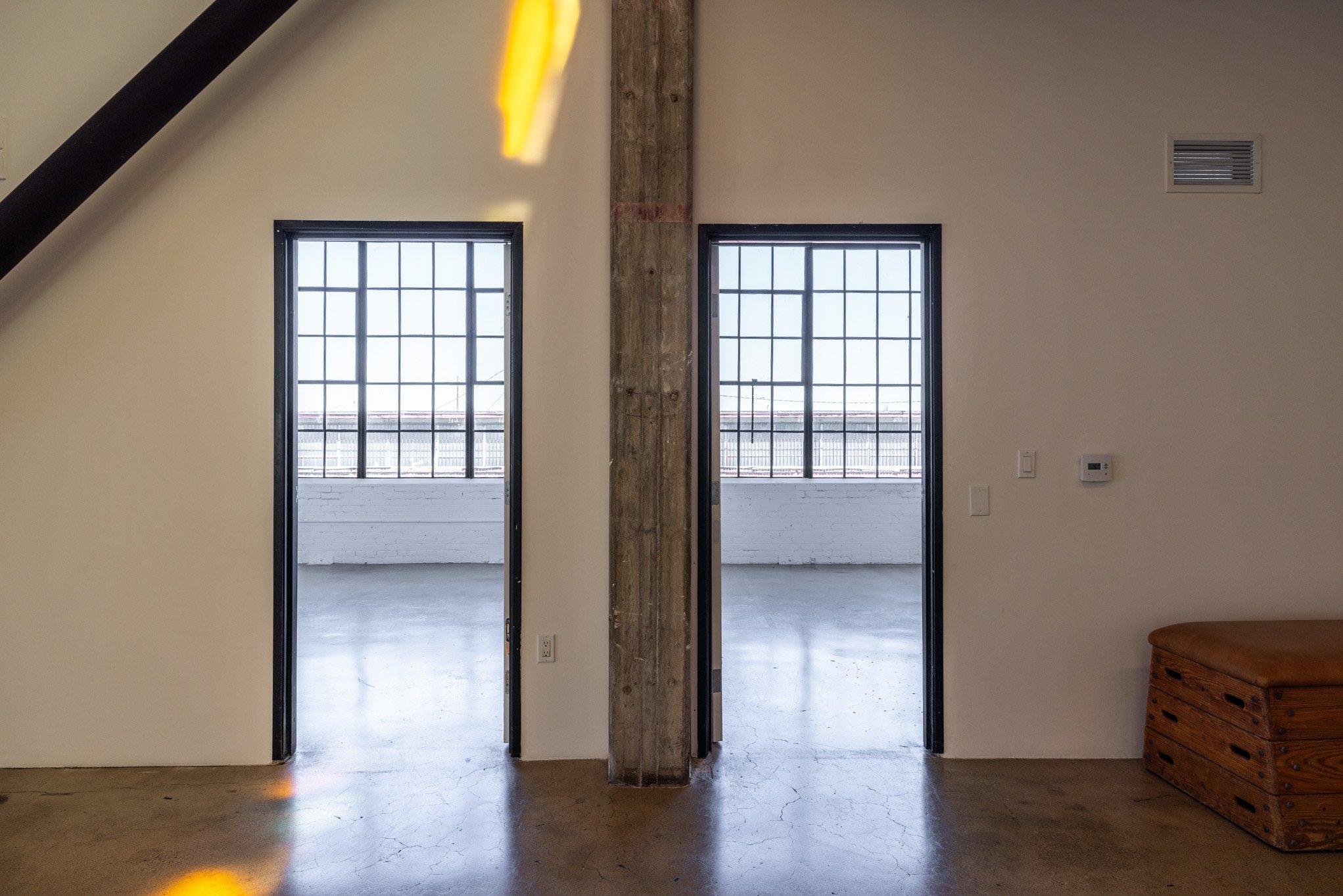 Interior of an industrial-style loft with two open doorways leading to a bright balcony, a visible concrete column, and a wooden bench on a polished concrete floor.