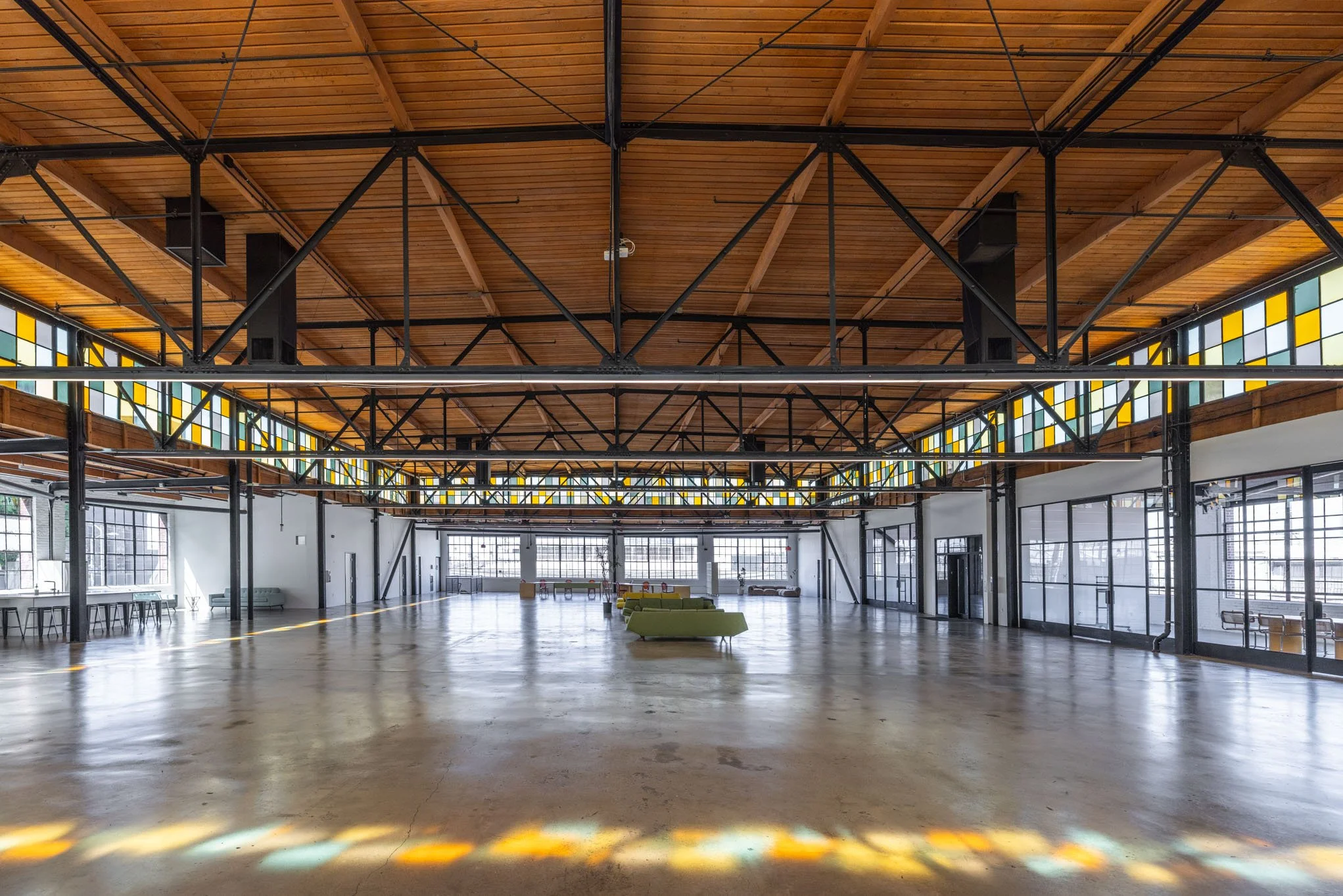Spacious industrial-style interior with polished concrete floors, large windows, exposed wooden ceiling, and black metal beams, featuring some green couches in the center.