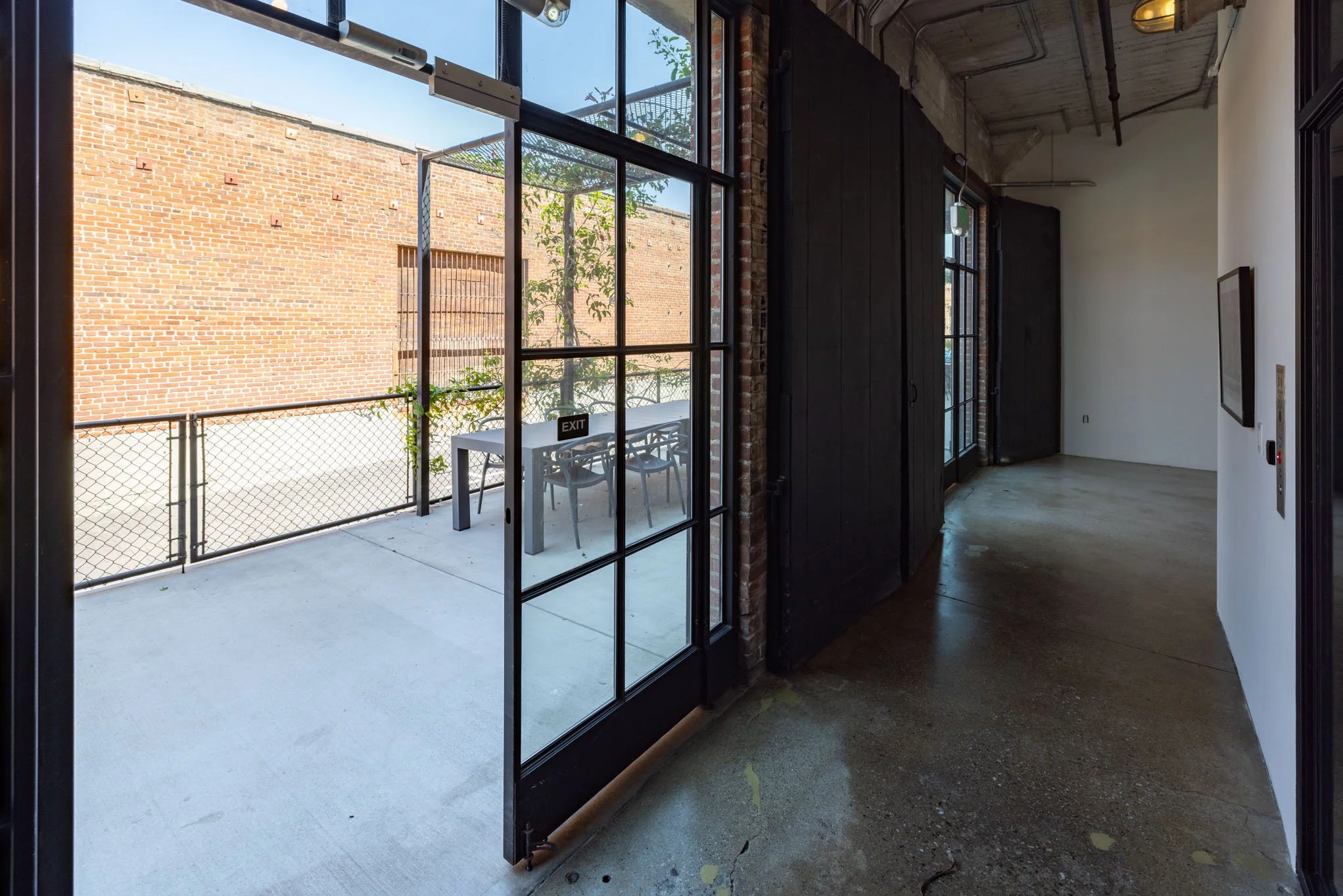 Indoor corridor with a glass door leading to a small outdoor patio with a table and chairs, brick wall in the background, and a tree visible outside.