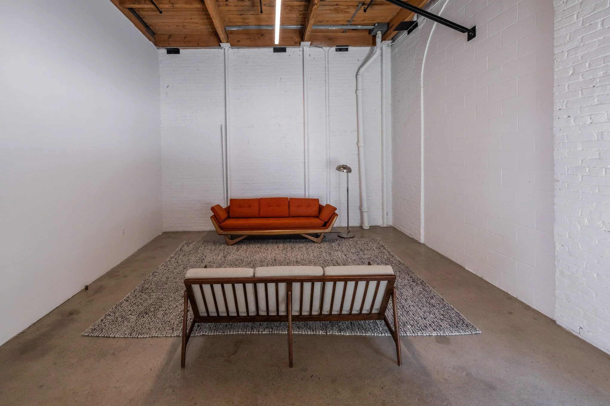 Minimalist living room corner with a mid-century modern orange sofa, a wooden bench with a white cushioned top, a gray area rug, a tall floor lamp, white brick and painted walls, and a wooden ceiling with a linear LED light.