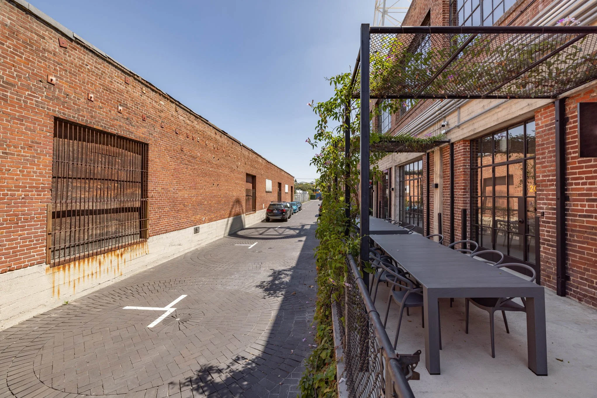 Urban alleyway with red brick walls, parked cars, a large metal barred window, and outdoor seating with a table and chairs