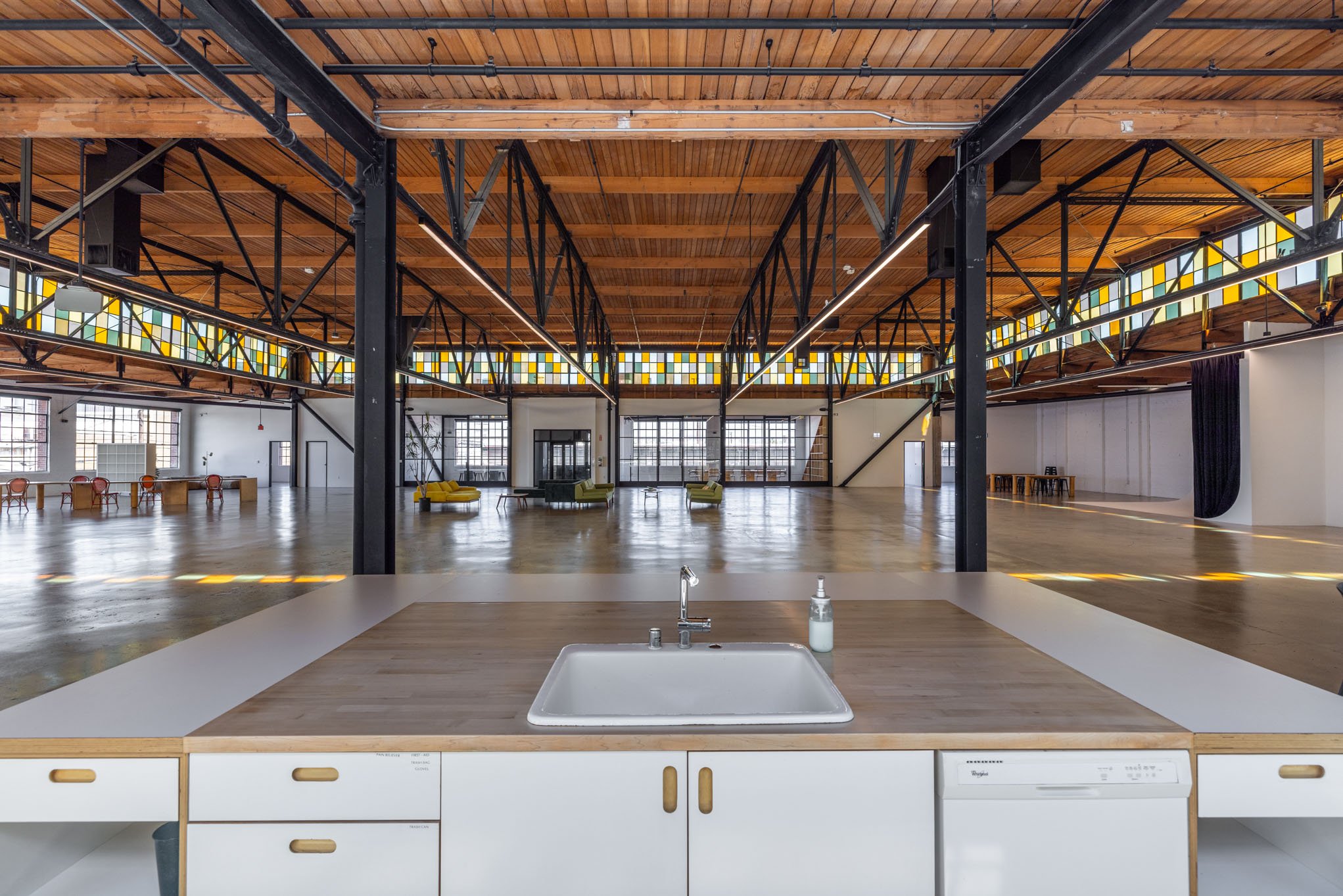 Open, spacious industrial-style loft with high wooden ceilings, exposed black metal beams, large windows, and colorful seating areas, viewed from the kitchen counter with sink and soap dispenser.
