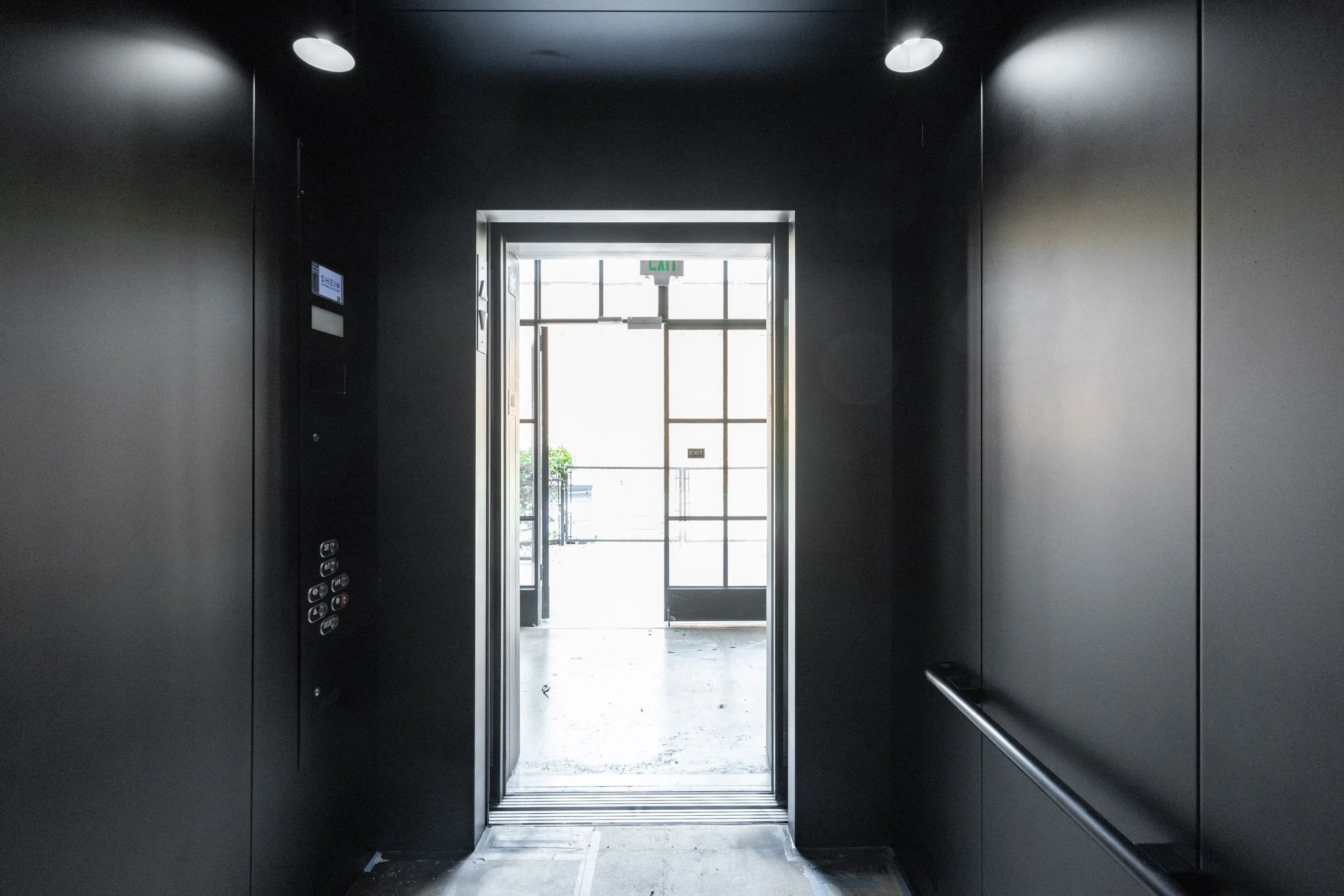 Interior view of an elevator with black walls, illuminated ceiling lights, and open door leading outside to a bright exterior with a green plant.