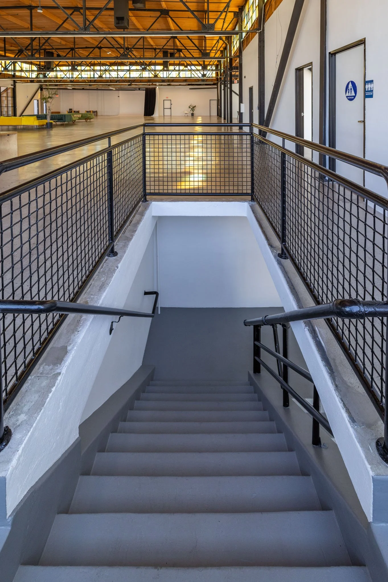 Indoor staircase with black handrails leading down, metal grid railing, view from top, in a modern industrial-style building with exposed beams and large windows.