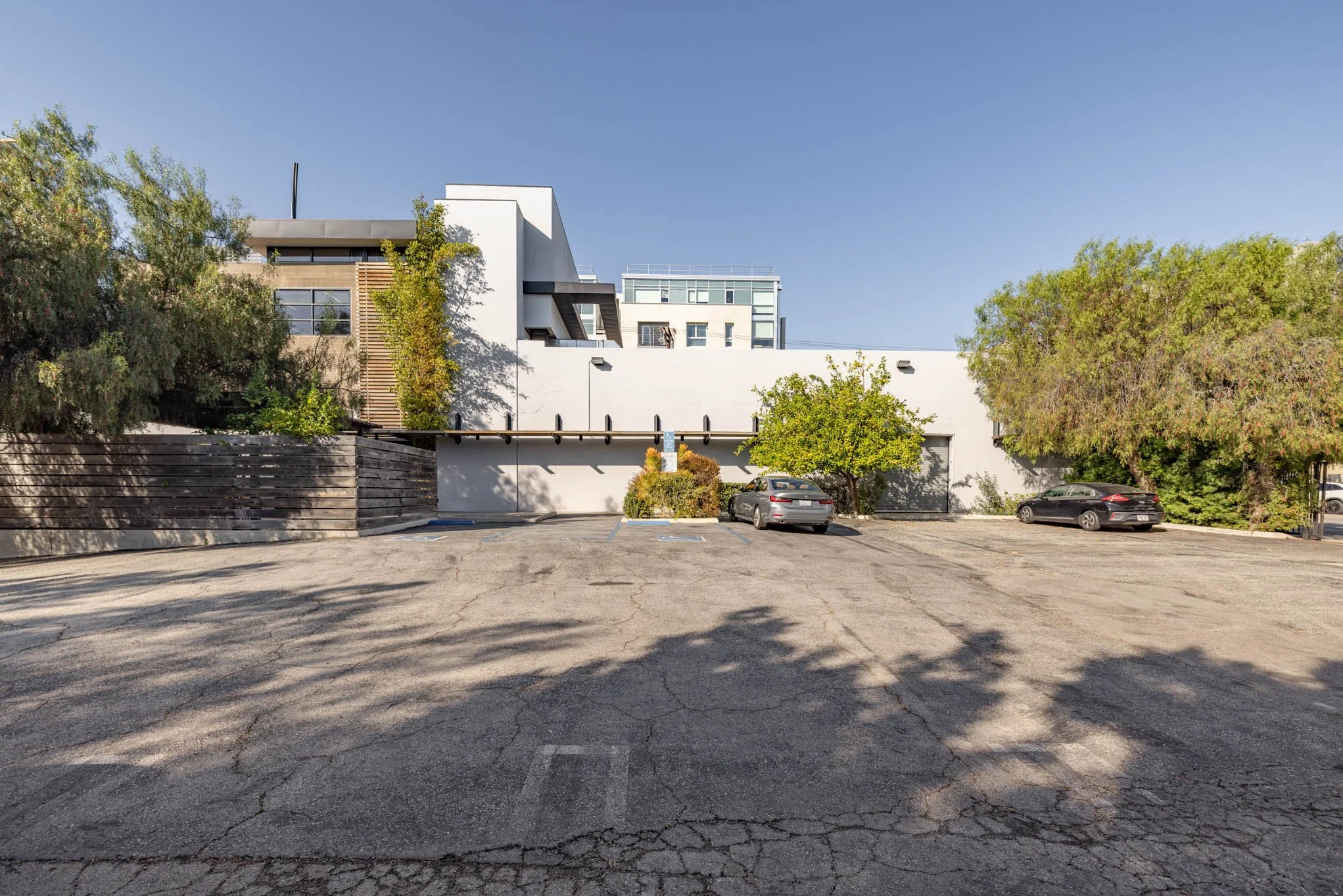 Empty parking lot with three parked cars, trees, and a modern building in the background under a clear blue sky.