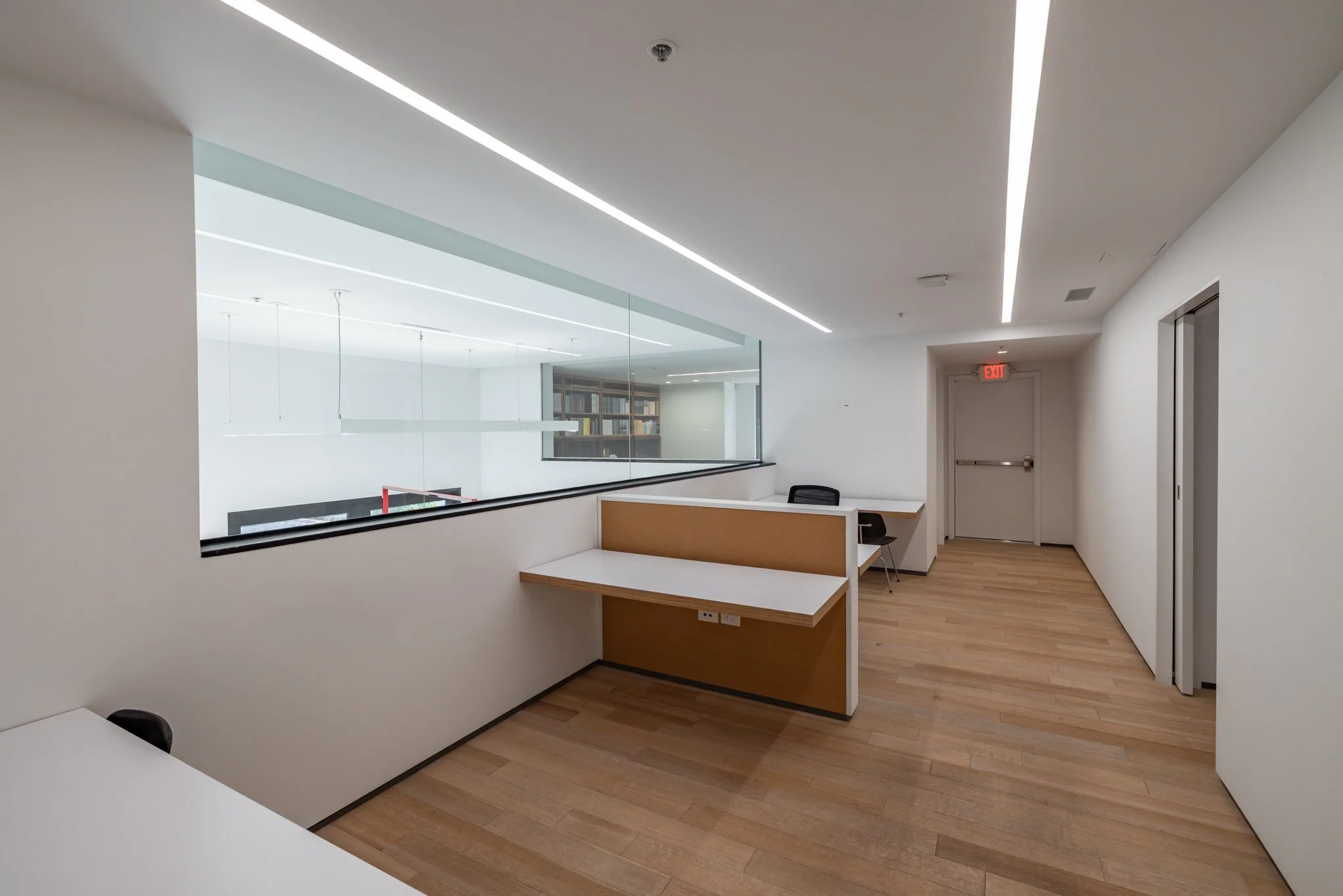 Empty Office corridor with wooden flooring, white walls, and ceiling. There is a desk with a brown divider on the left, a small table with a black chair against the wall, and a glass window looking into another room with bookshelves. An exit door wit