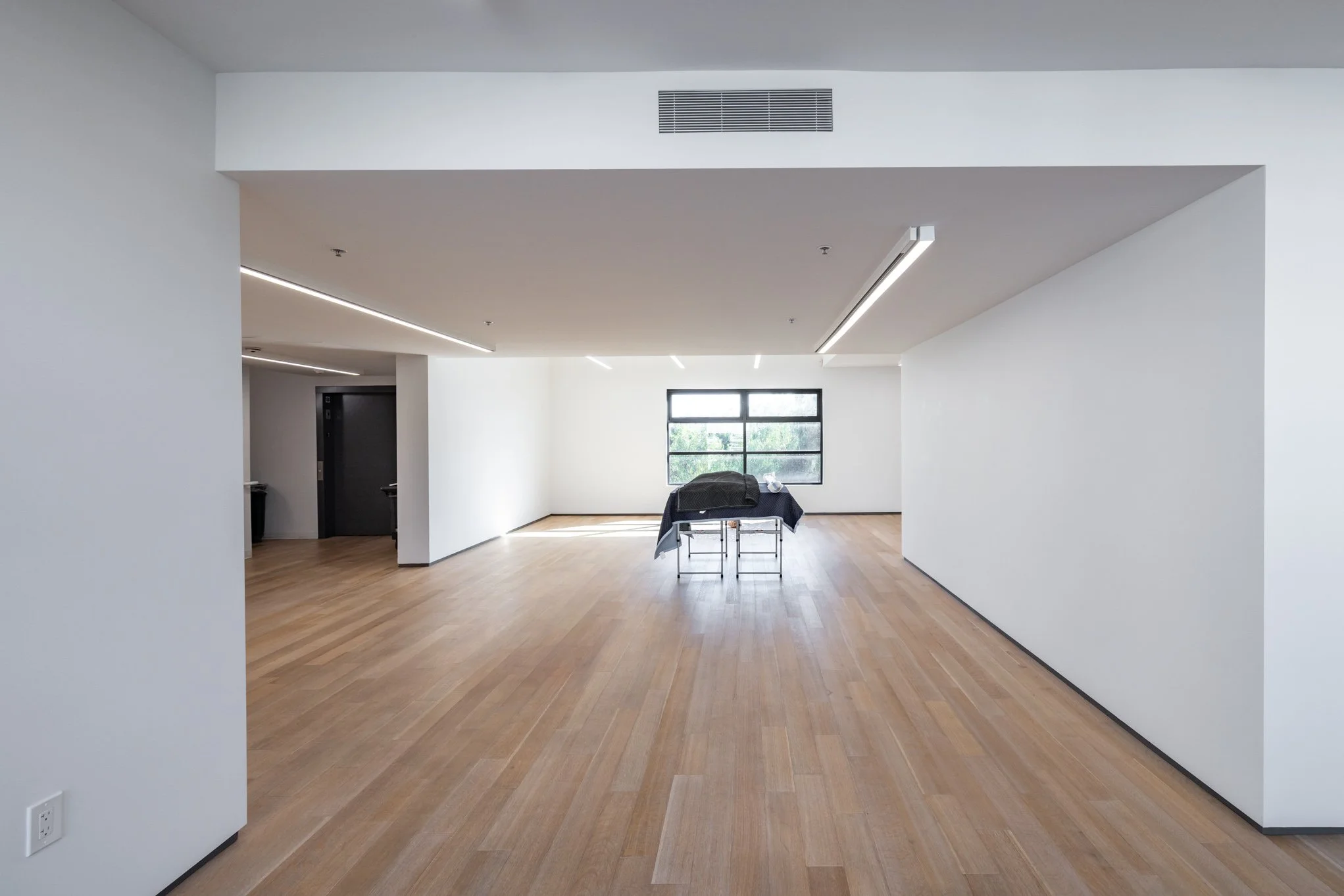 Empty modern apartment with light wood flooring, white walls, a large window, and a table covered with a cloth in the center.