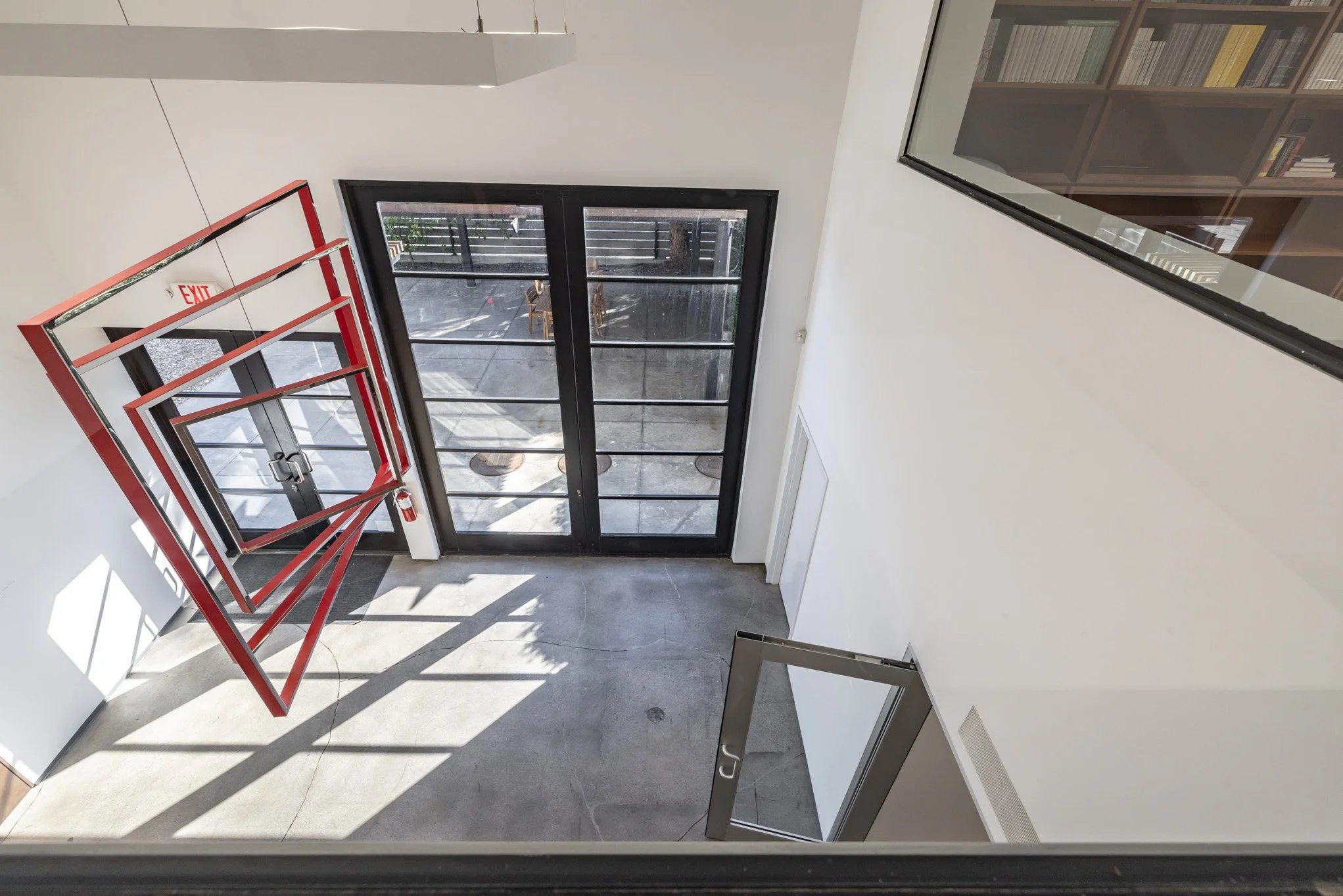 Inside view of a modern building entrance with black-framed glass double doors, a large glass window, a red fire exit sign and gate, concrete floor, and part of a second floor with a bookshelf visible through a window.