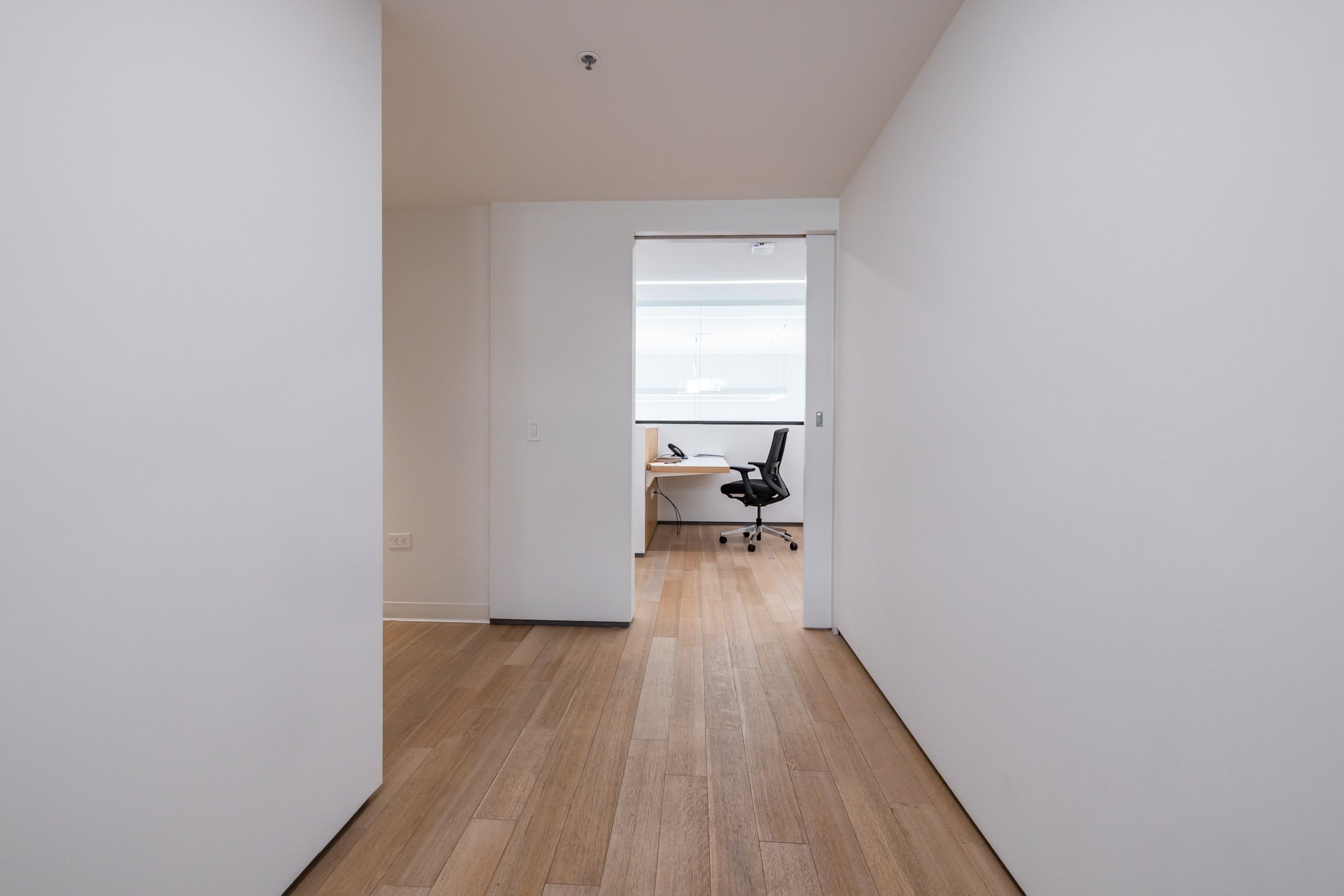 Empty hallway leading to a bright office space with a black office chair, desk, and window.