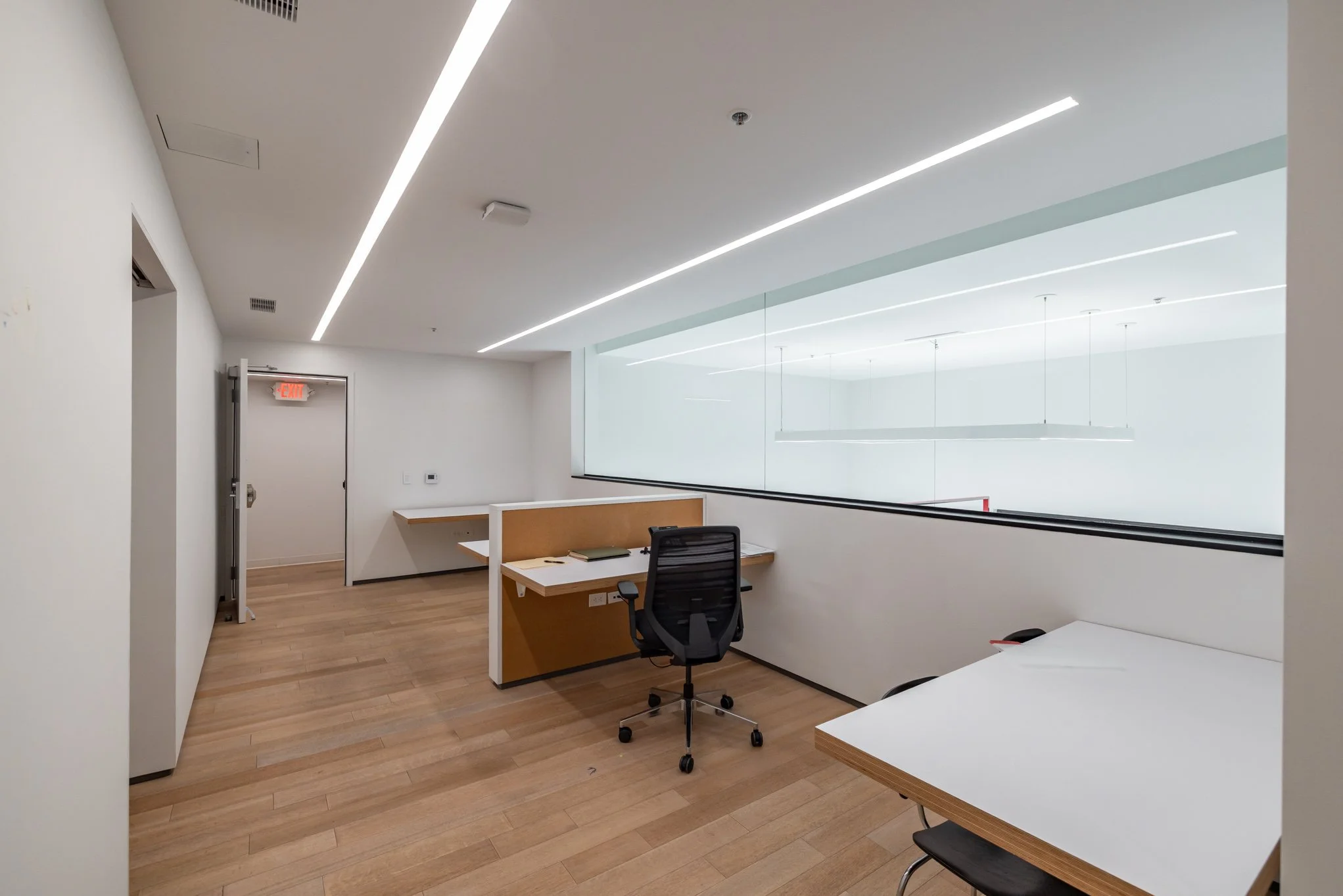 Modern office space with white walls, wood flooring, a desk with a black office chair, and large windows.