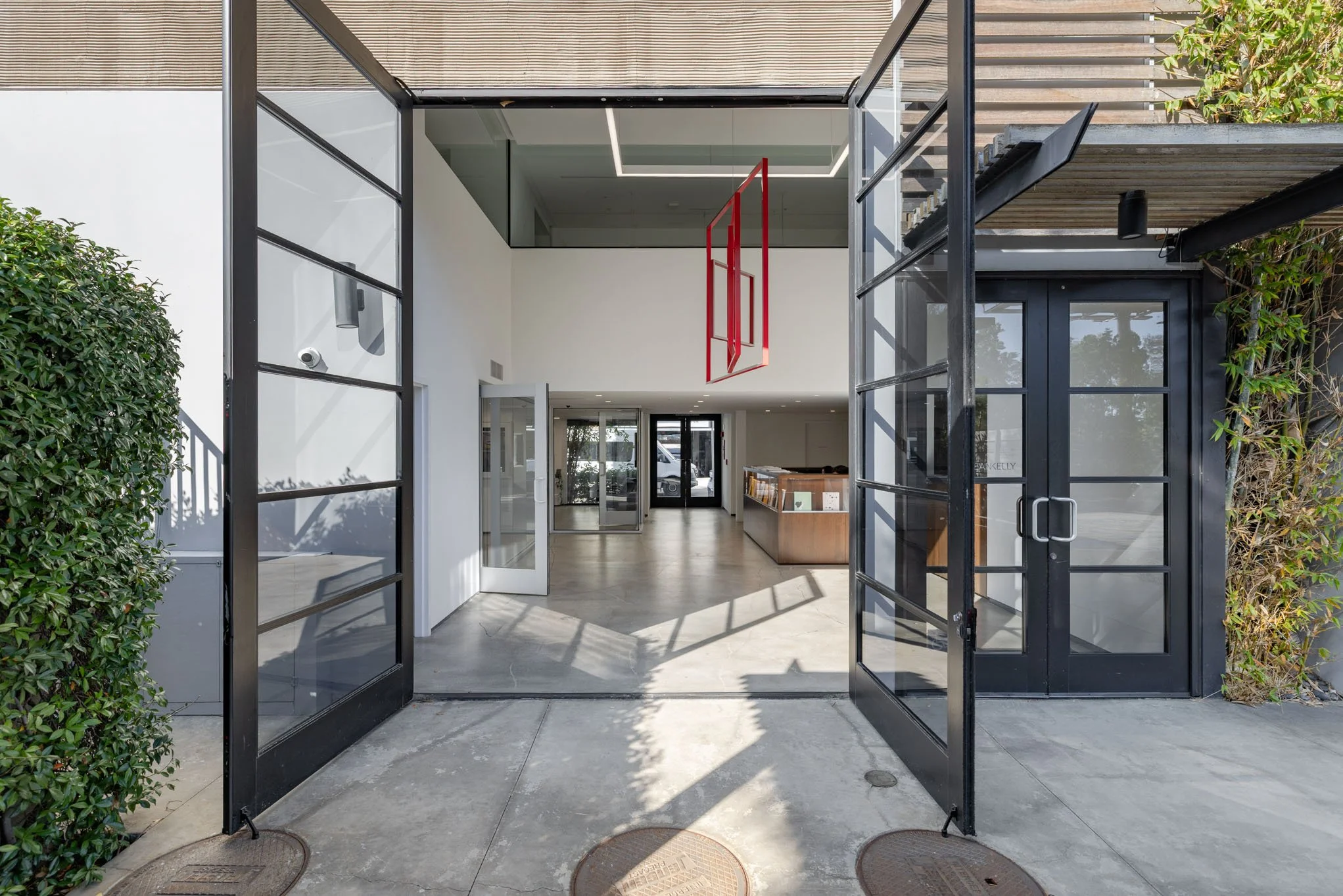 Open black metal framed glass doors leading into a modern building lobby with white walls, a reception desk, a red geometric art installation hanging overhead, and natural sunlight streaming inside.