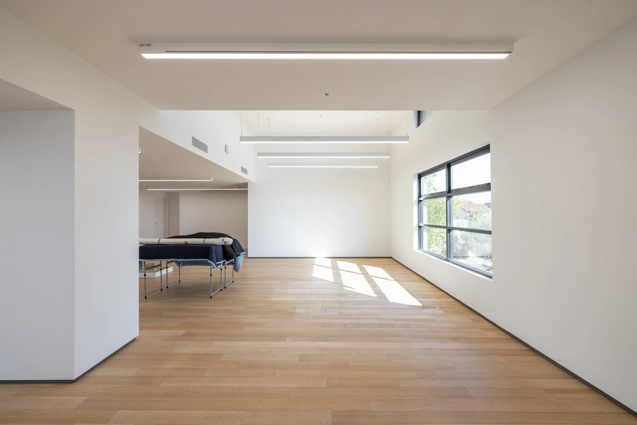 Empty room with large windows, light wooden flooring, white walls, and modern ceiling lights, with a table and some items in the corner.