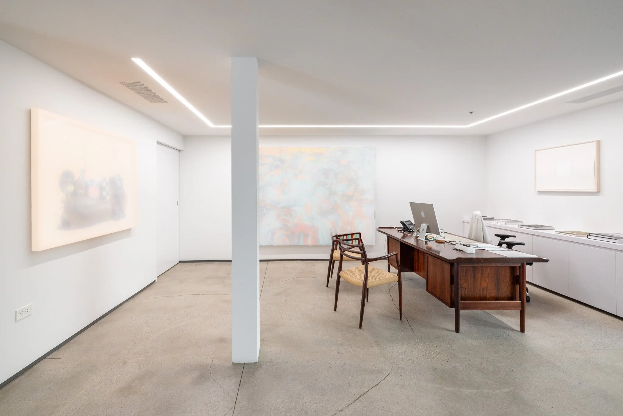 Minimalist office with white walls, abstract artwork, a wooden desk with a computer, chairs, and a white storage unit under a ceiling with integrated lighting.