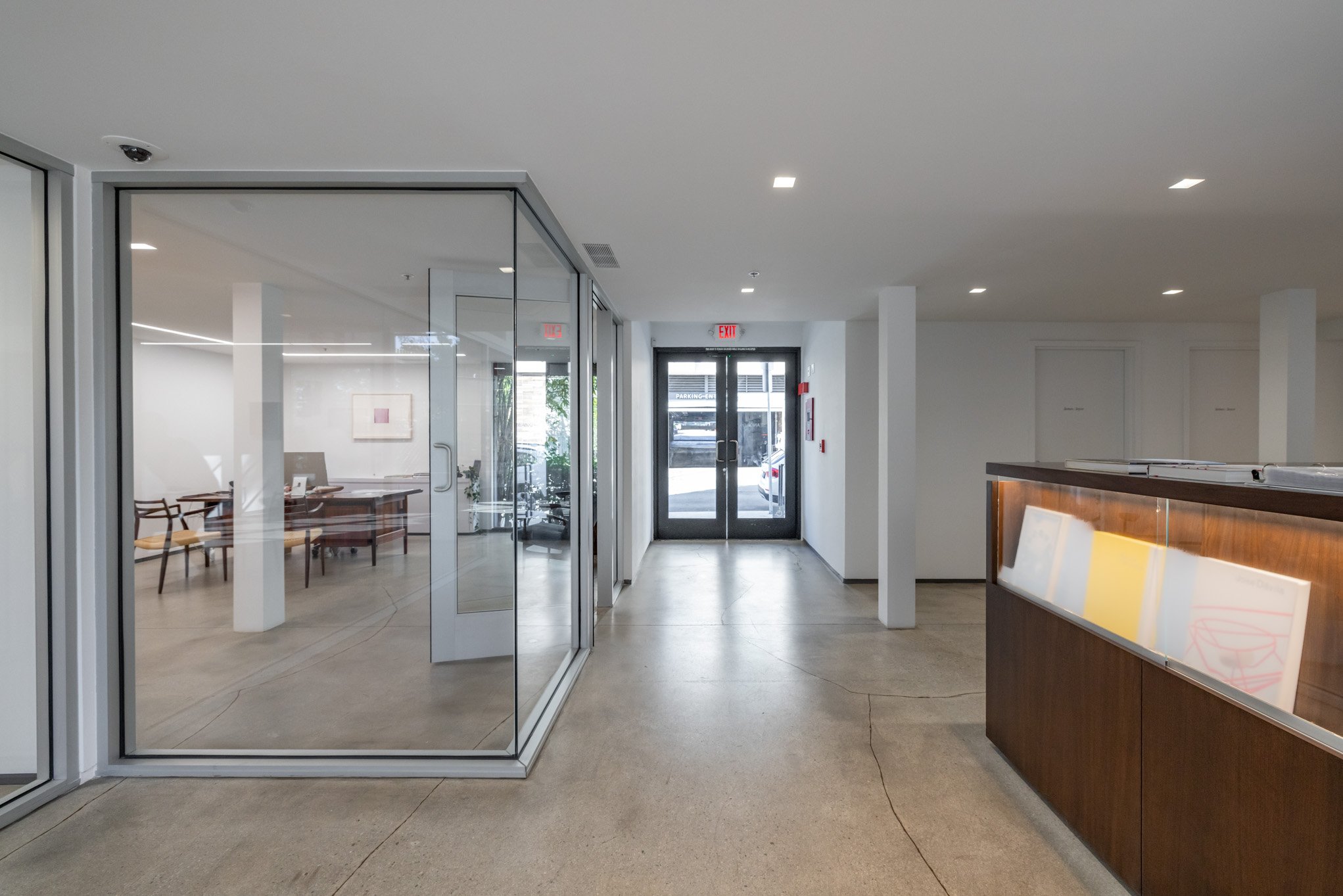 Interior view of a modern building with a glass-enclosed office on the left, a reception desk on the right, and a glass door at the end leading outside, with a small lobby area.