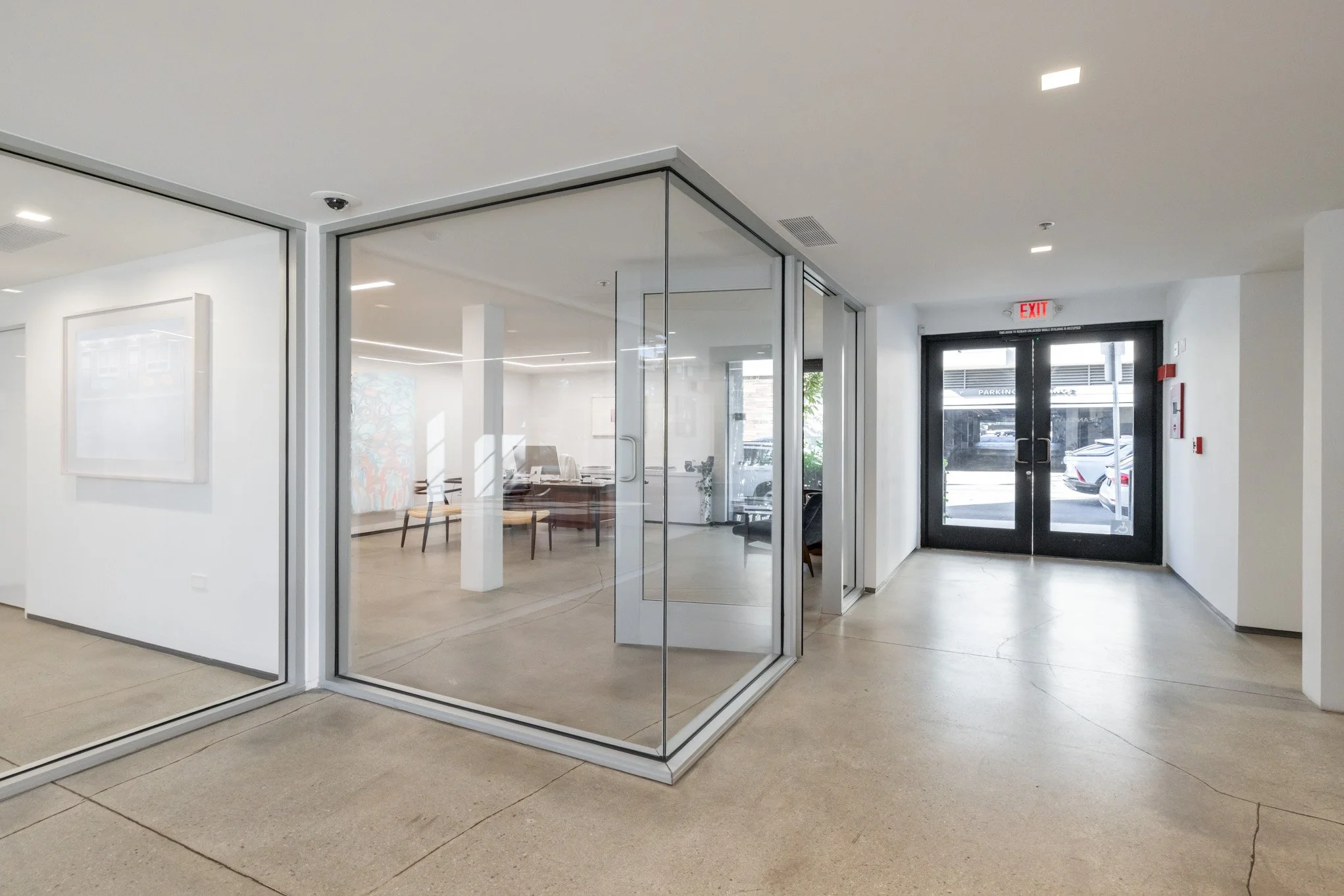 Interior shot of a modern office building entrance with glass walls, a dark door with an exit sign, and a parking lot visible outside.