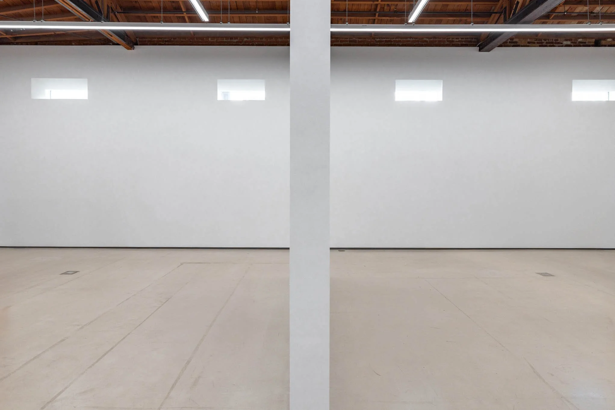 Empty room with white walls, small rectangular windows, and a concrete pillar in the center. Exposed wooden ceiling beams with industrial lighting.