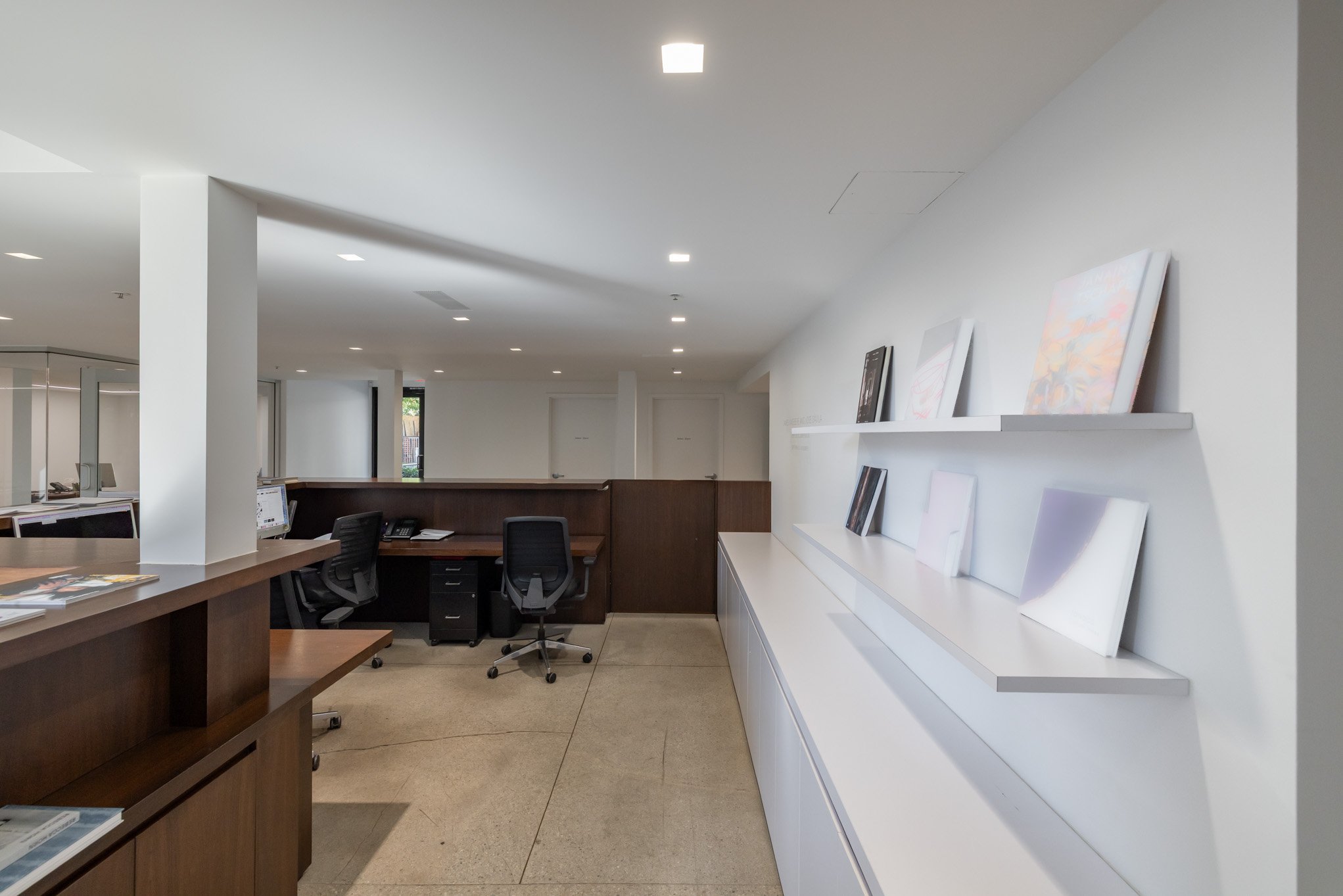 An office reception area with a white wall on the right displaying paintings on a white shelf. On the left, there are dark wooden desks with black office chairs, some with phones and computers. The floor is beige, and the ceiling has multiple small c
