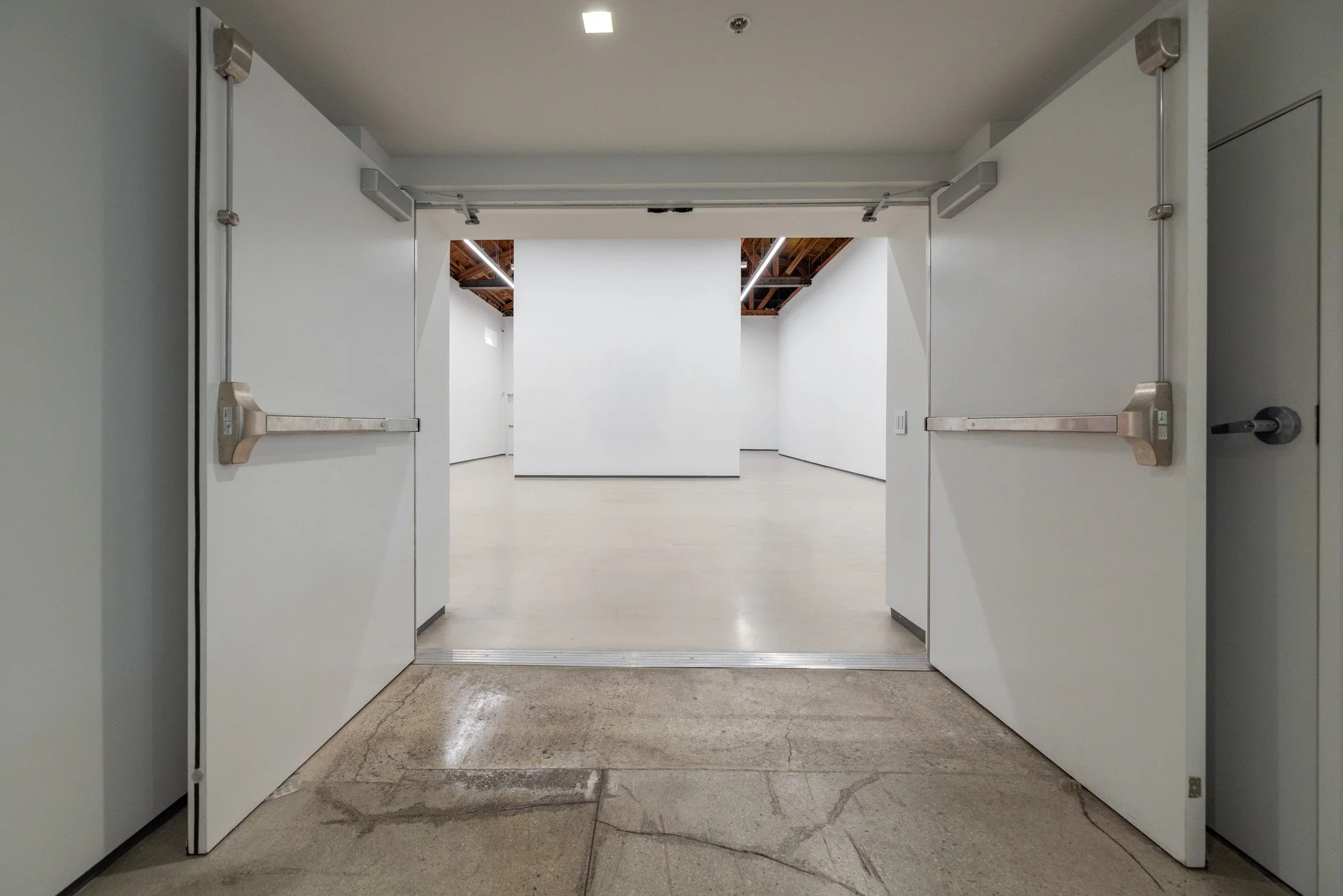 Open white sliding doors leading into an empty art gallery with white walls and a wooden ceiling.
