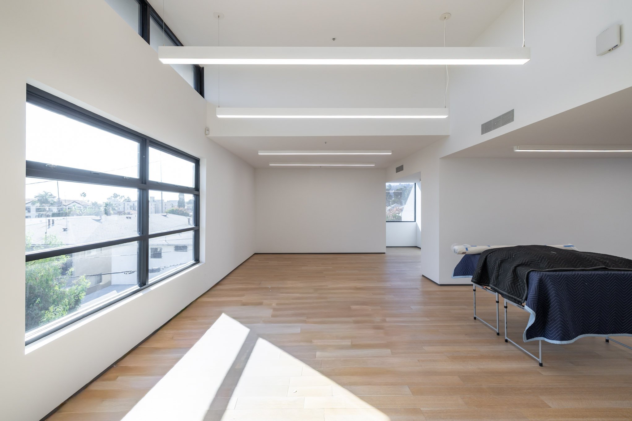 Empty room with large windows, wooden flooring, and a covered piece of furniture on the right.