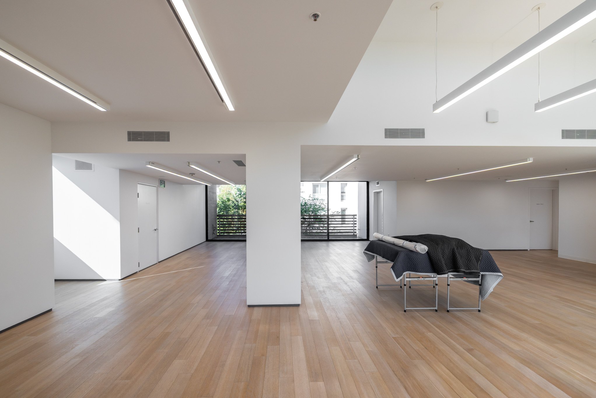 An empty modern interior room with wooden floors, white walls, and ceiling with modern strip lighting. There are large windows and doors leading to balconies, and some furniture covered with a black cloth.