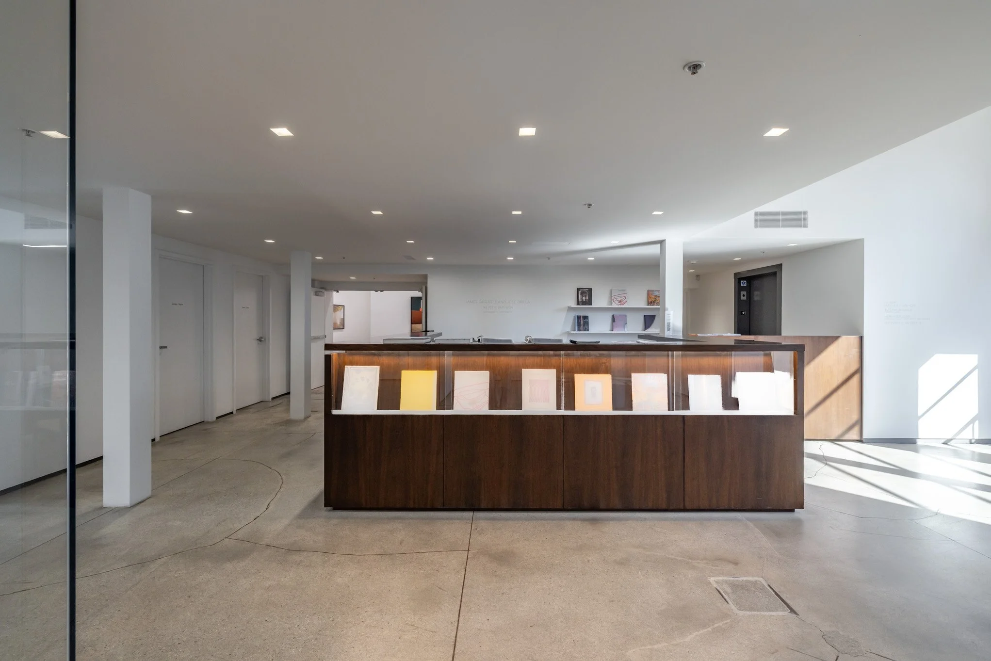 An interior space with a reception desk made of dark wood, display cases with framed pictures, white walls, and a white ceiling with recessed lighting. There are doors on the left and some artwork on the far wall.
