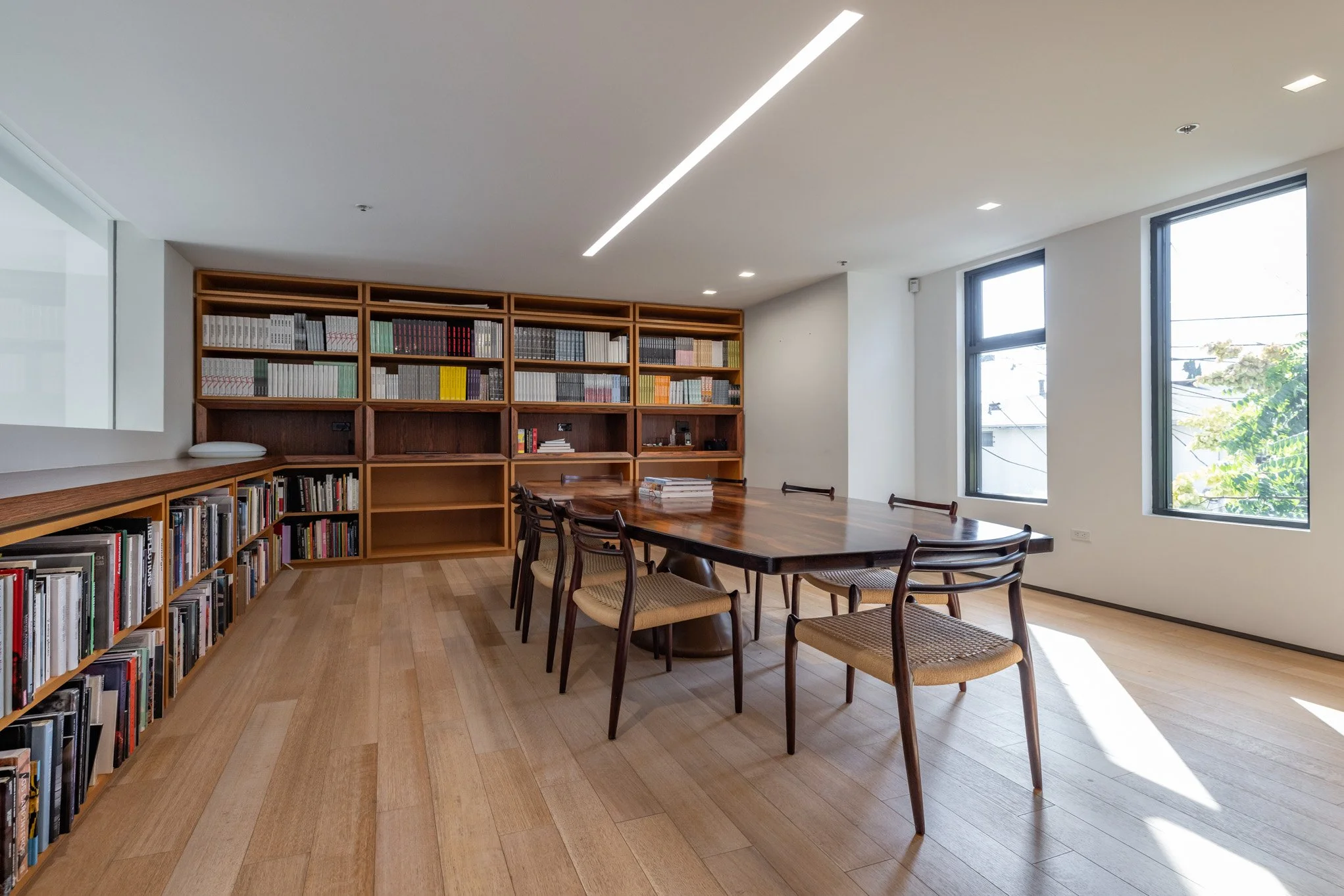 A modern, well-lit library or conference room featuring a large wooden table with six chairs, bookshelves filled with books along the back wall, and large windows letting in natural light.