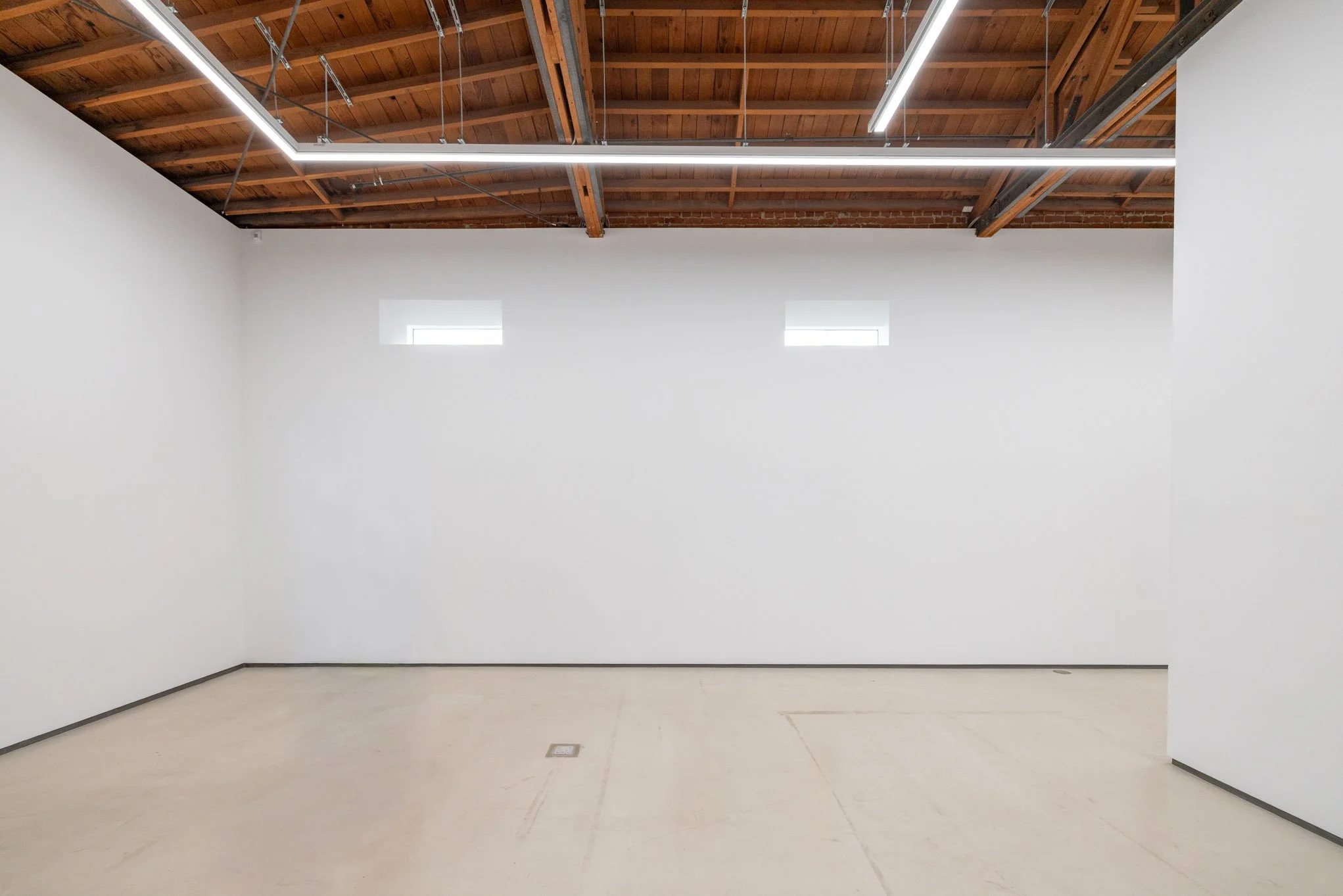 Empty art gallery with white walls, a wooden ceiling, and three small rectangular windows high on the wall. Fluorescent lighting fixtures are installed on the ceiling.