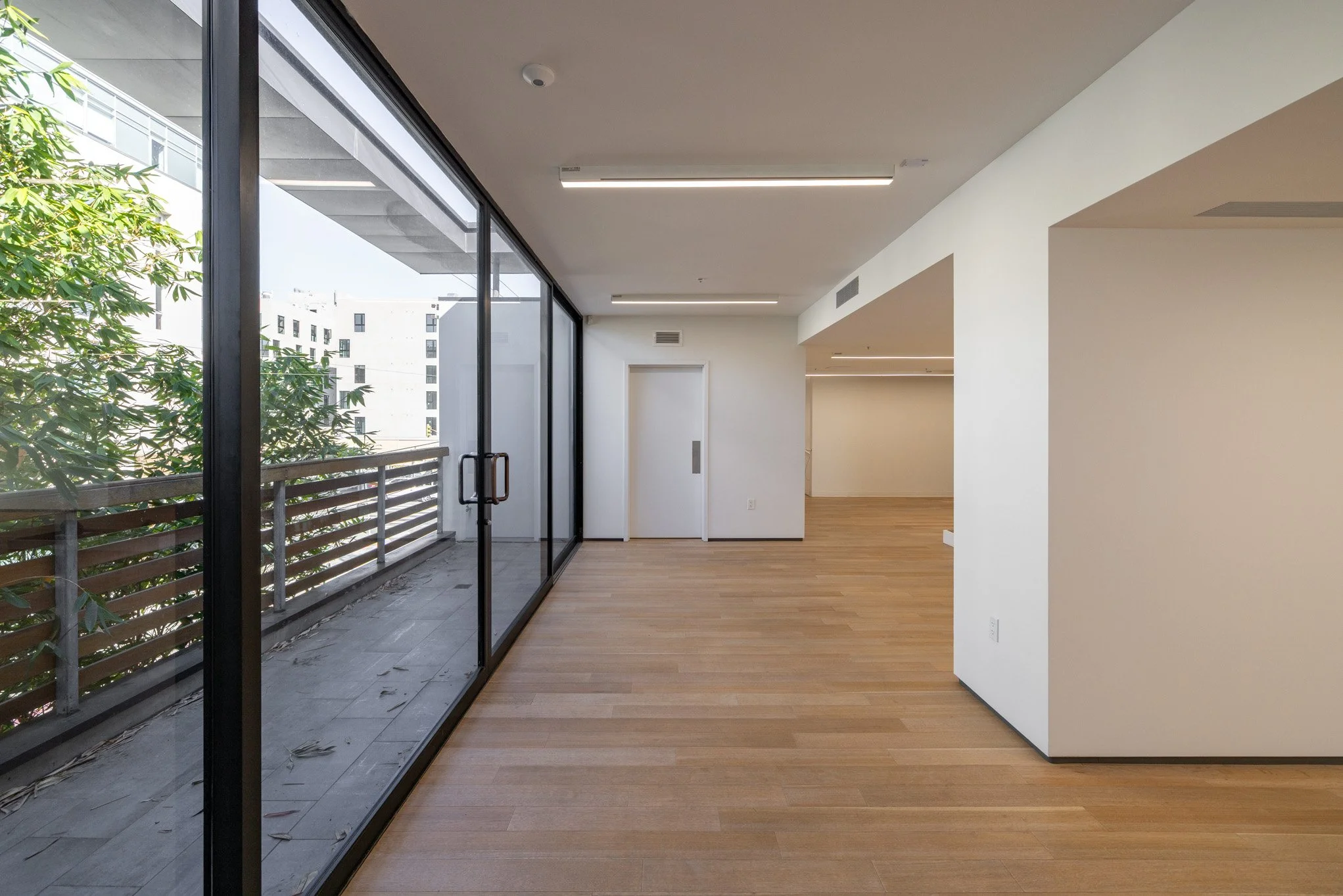 Empty modern apartment interior with large glass sliding door leading to balcony, hardwood floors, white walls, and ceiling lights.