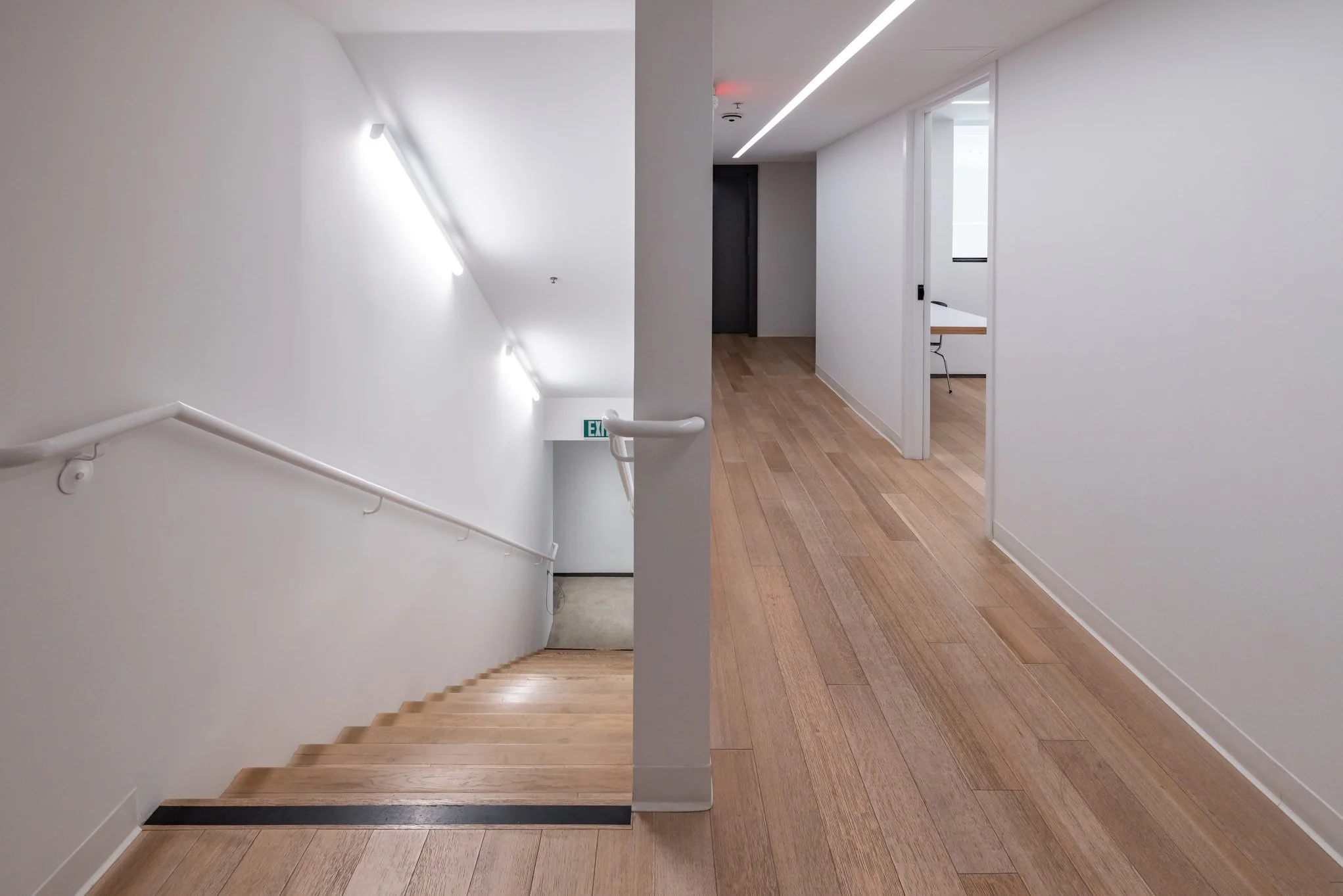 A modern, minimal staircase with wooden steps on the left, white walls, and a wooden floor on the right with a hallway leading to a door and a small office space visible in the background.