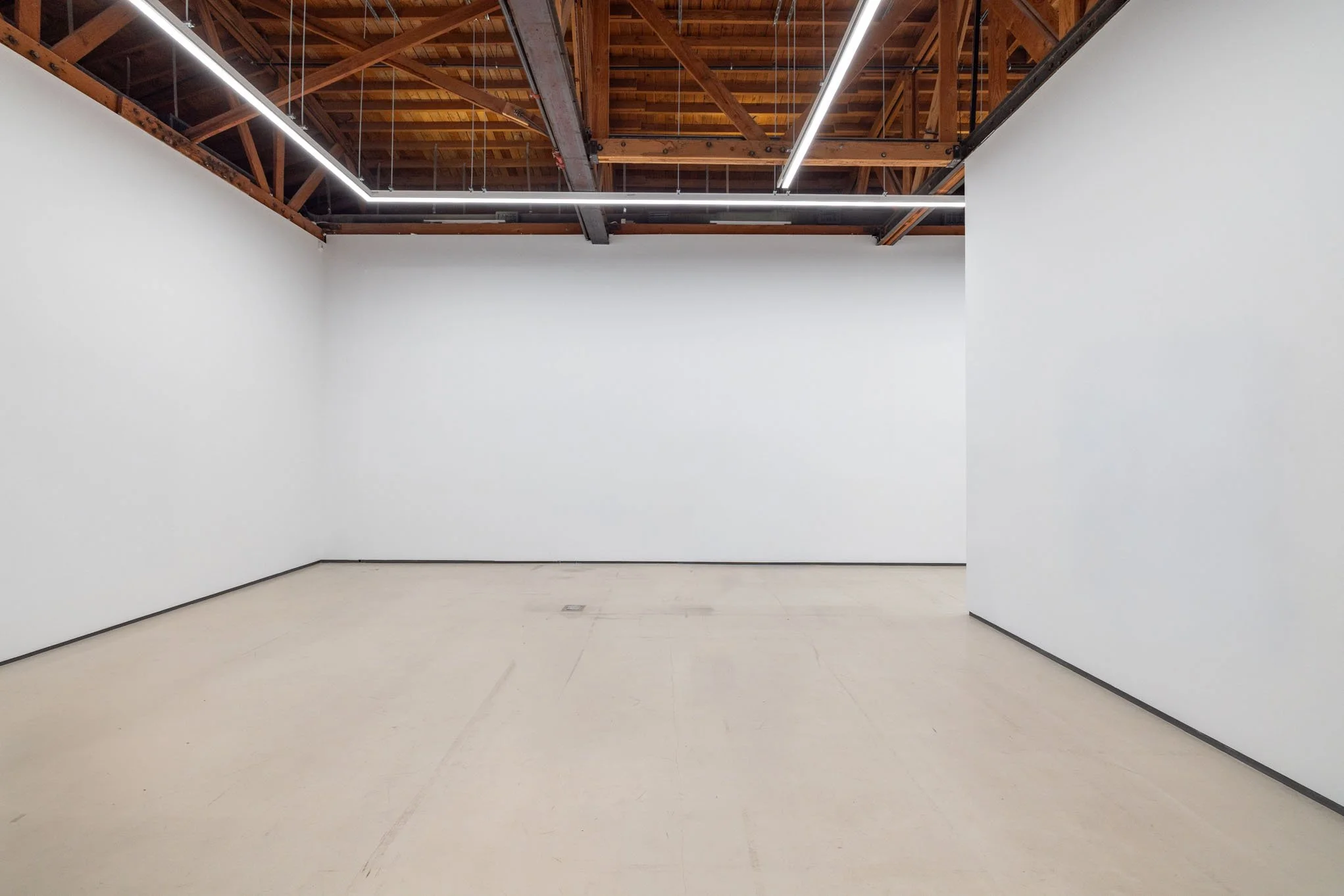 Empty art gallery with white walls, beige flooring, and exposed wooden ceiling with metal beams and linear lighting.
