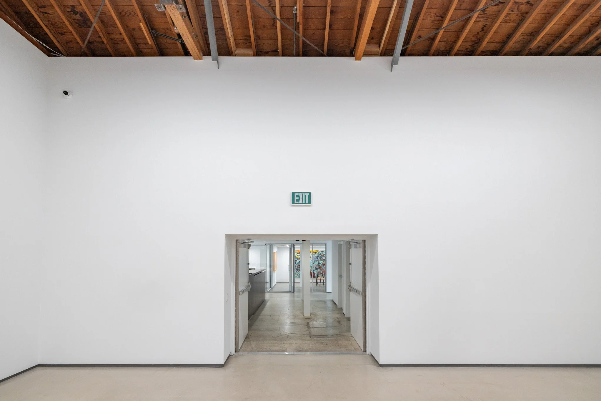 Empty white-walled room with high ceiling, wooden beams, and an open doorway leading to a hallway with artwork and chairs, and an illuminated green exit sign above the doorway.